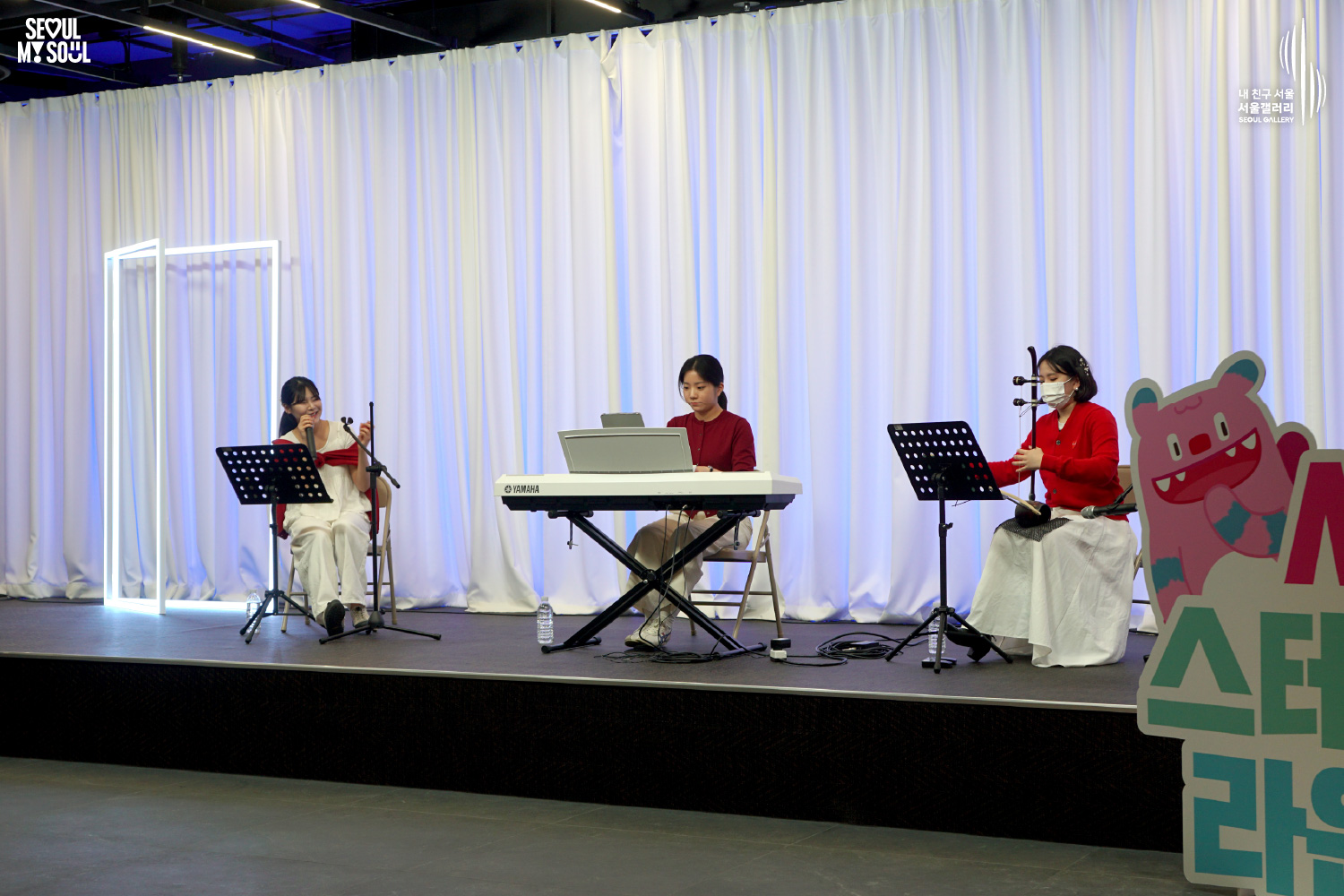 A female trio of two vocalists and a Yamaha keyboard player performs on a white-curtained stage at Seoul Gallery, with "SEOUL MY SOUL" and "Seoul Gallery" logos visible.