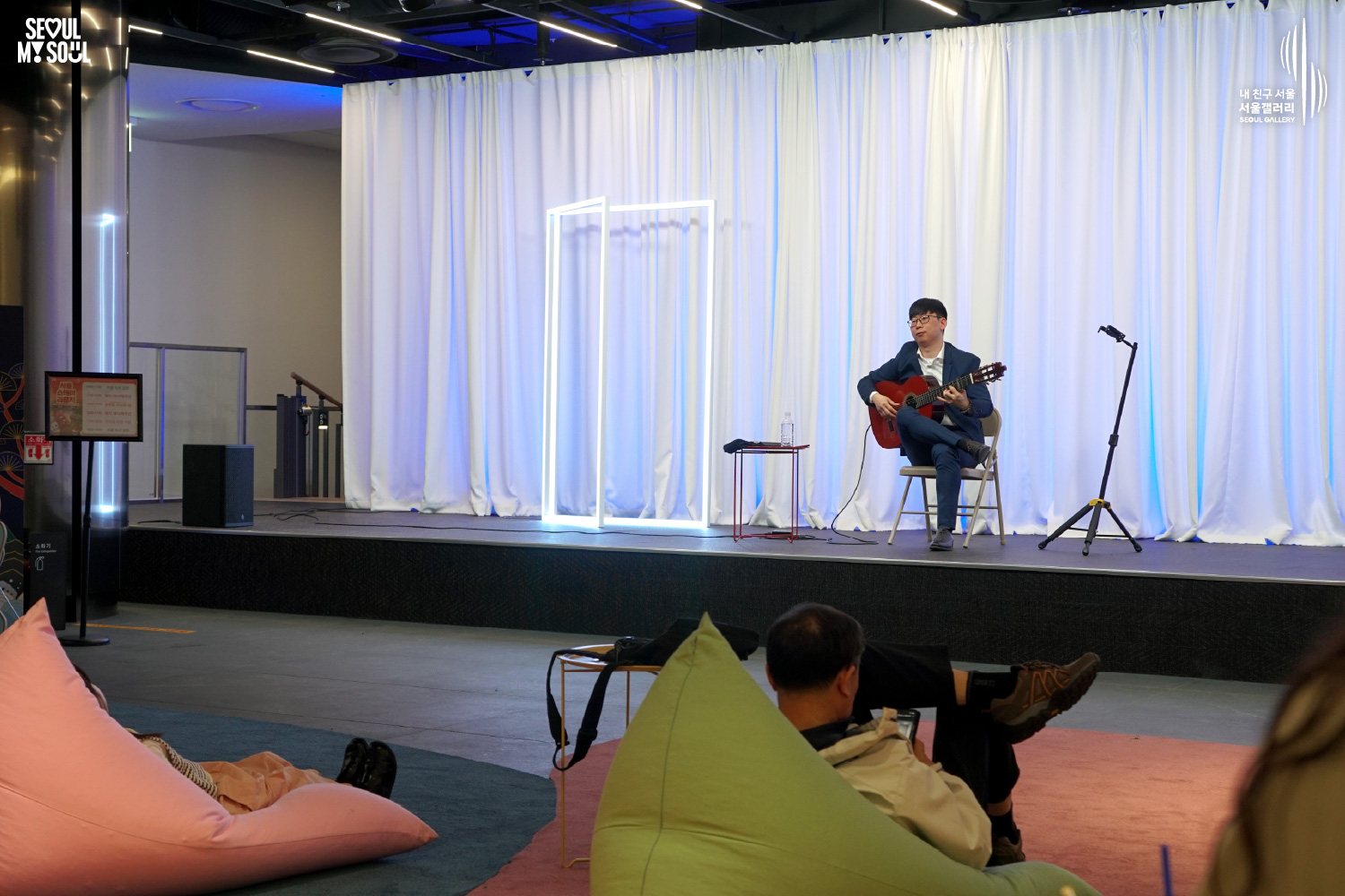 A classical guitarist in a navy suit performs seated on a blue-lit stage at Seoul Gallery while audience members relax on beanbag chairs, with "SEOUL MY SOUL" and "Seoul Gallery" logos visible.