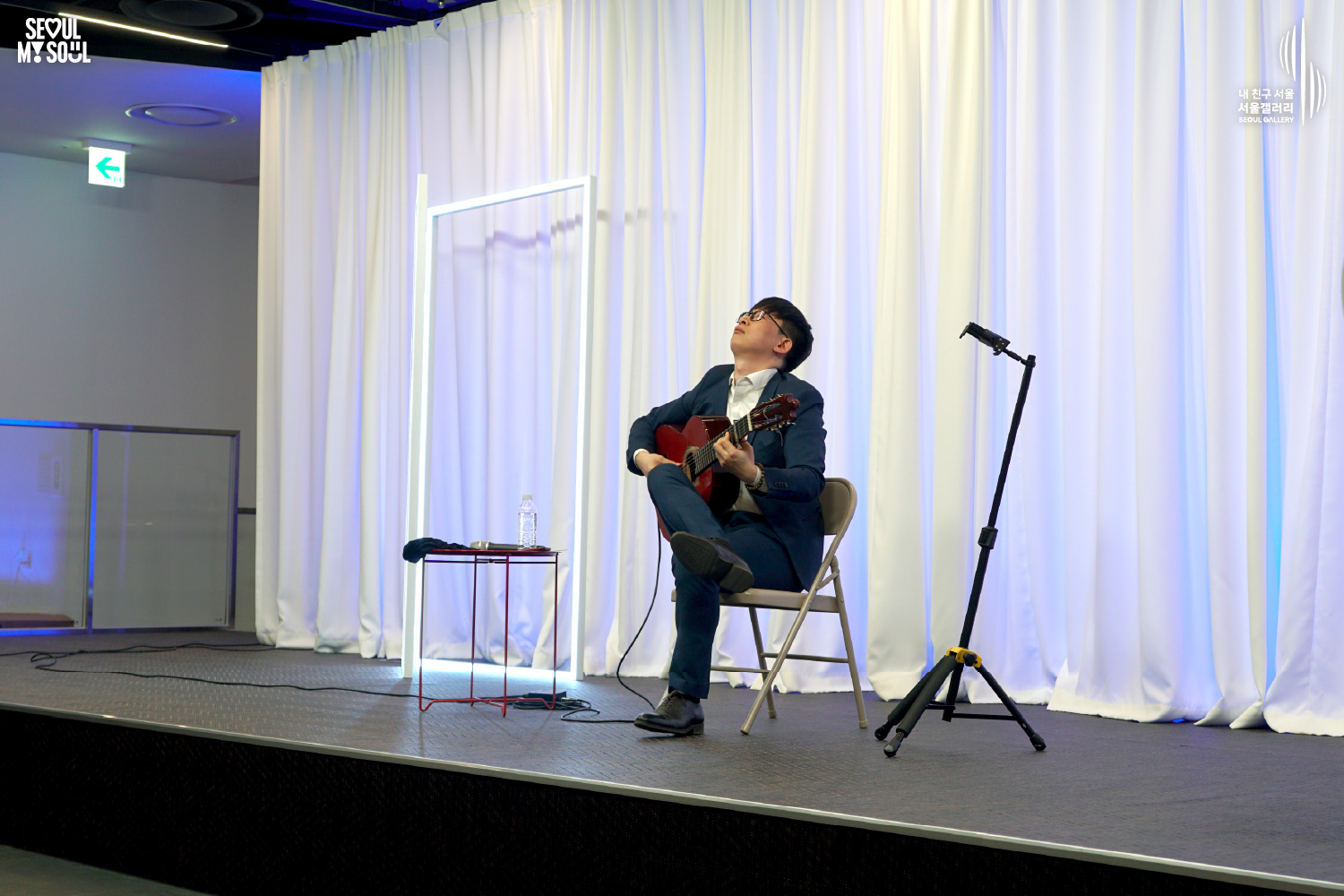 A classical guitarist in a navy suit performs seated on a blue-lit stage with white curtains at Seoul Gallery, with "SEOUL MY SOUL" and "Seoul Gallery" logos visible.