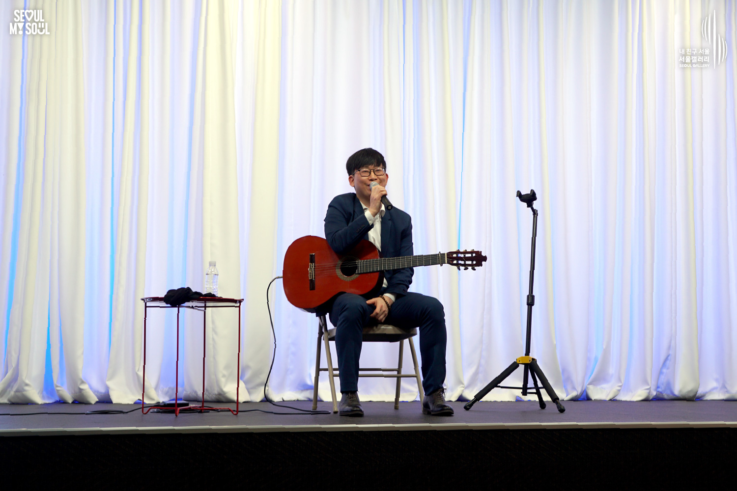 A classical guitarist in a navy suit speaks into a microphone while seated with a guitar on a blue-lit stage at Seoul Gallery, with "SEOUL MY SOUL" and "Seoul Gallery" logos visible.