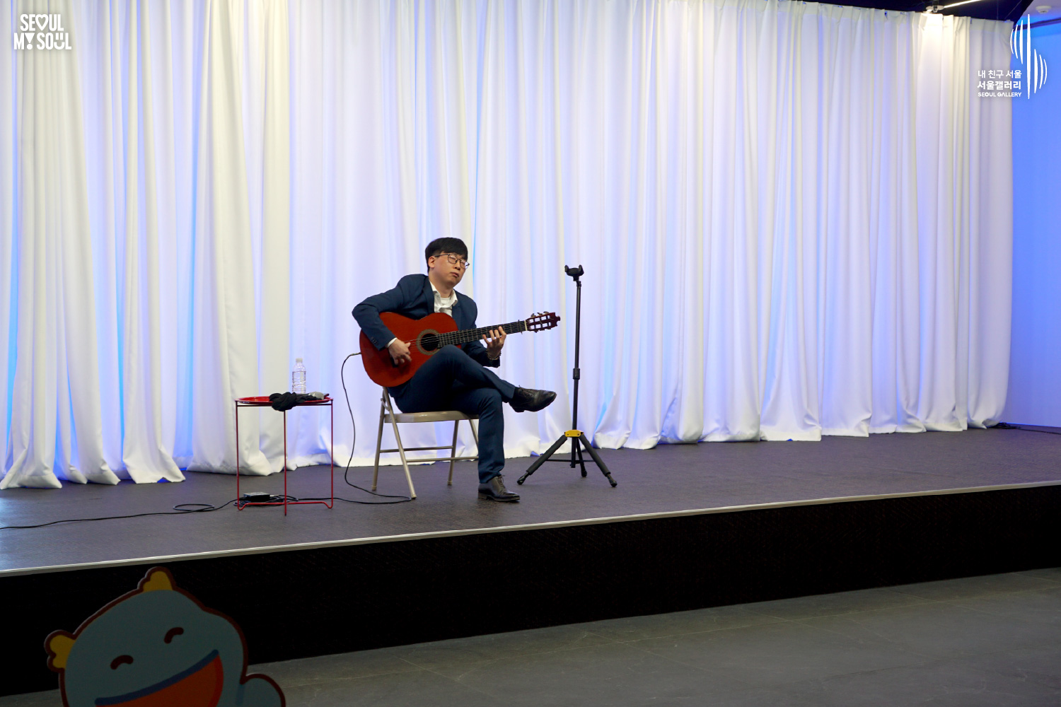 A classical guitarist in a navy suit performs seated on a blue-lit stage with white curtains at Seoul Gallery, with "SEOUL MY SOUL" and "Seoul Gallery" logos visible.