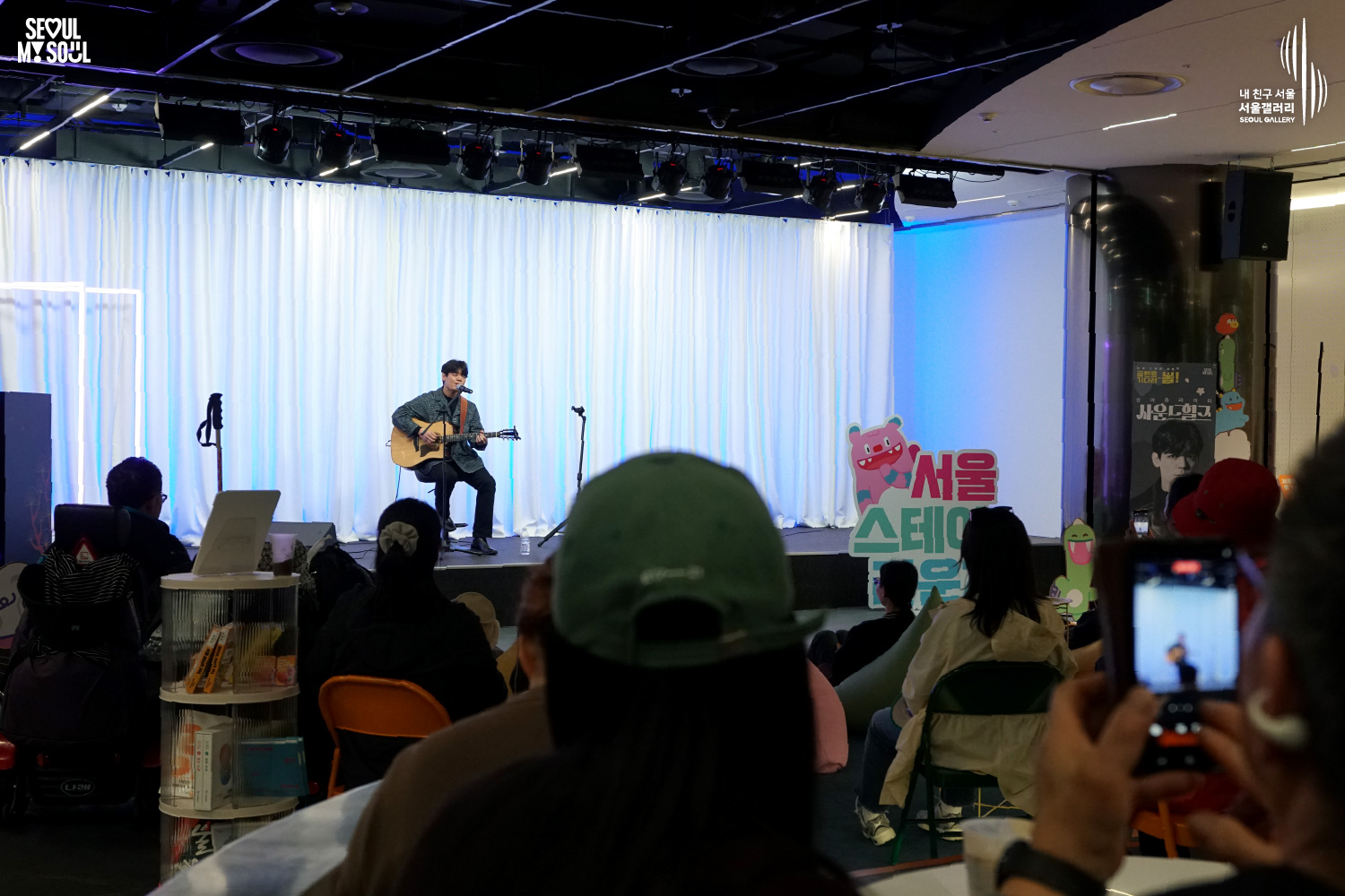 A solo acoustic guitarist performs seated on stage at Seoul Gallery's Seoul Stage event before an audience, with "SEOUL MY SOUL" and "Seoul Gallery" logos visible.