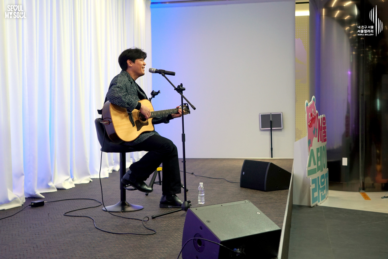 A solo acoustic guitarist performs seated on a stool at Seoul Gallery's Seoul Stage event, with "SEOUL MY SOUL" and "Seoul Gallery" logos visible.