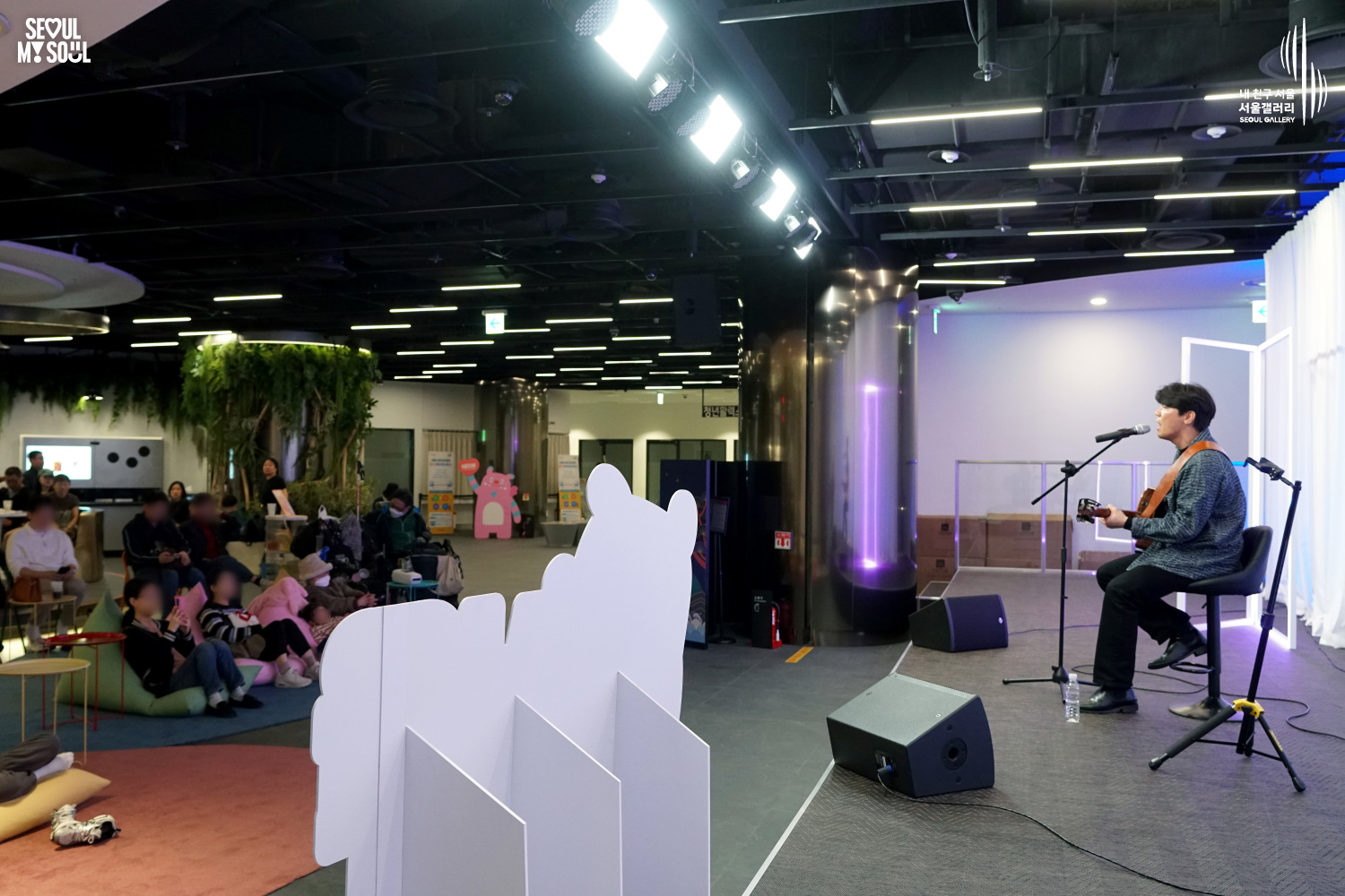 A solo acoustic guitarist performs seated at Seoul Gallery while audience members relax on beanbags and lounge chairs, with "SEOUL MY SOUL" and "Seoul Gallery" logos visible.