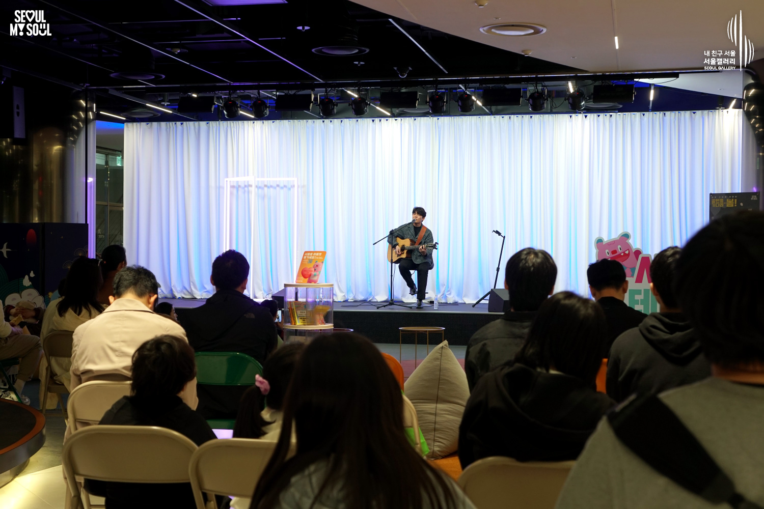 A solo acoustic guitarist performs seated at Seoul Gallery's Seoul Stage event before a seated audience, with "SEOUL MY SOUL" and "Seoul Gallery" logos visible.