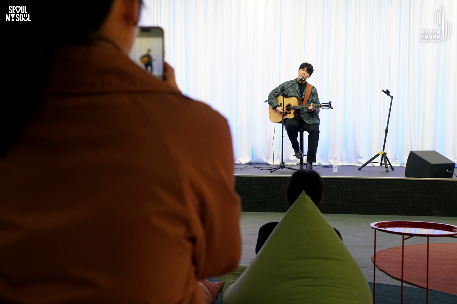 An audience member films a seated solo acoustic guitarist's performance on a stage with white curtains at Seoul Gallery, with "SEOUL MY SOUL" and "Seoul Gallery" logos visible.