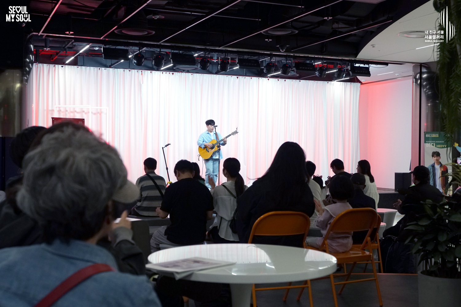 Singer-songwriter HEDYY performs with an acoustic guitar at Seoul Gallery's Lunch Stage event before a seated audience, with "SEOUL MY SOUL" and "Seoul Gallery" logos visible.