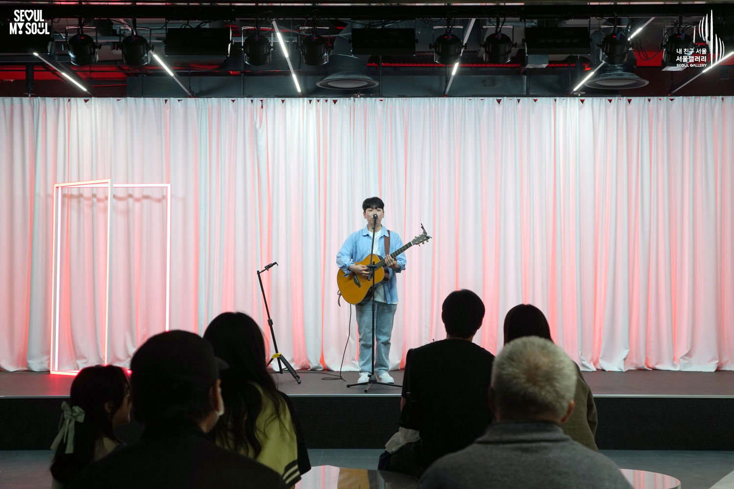 A solo acoustic guitarist sings on a pink-lit stage with white curtains at Seoul Gallery before a seated audience, with "SEOUL MY SOUL" and "Seoul Gallery" logos visible.