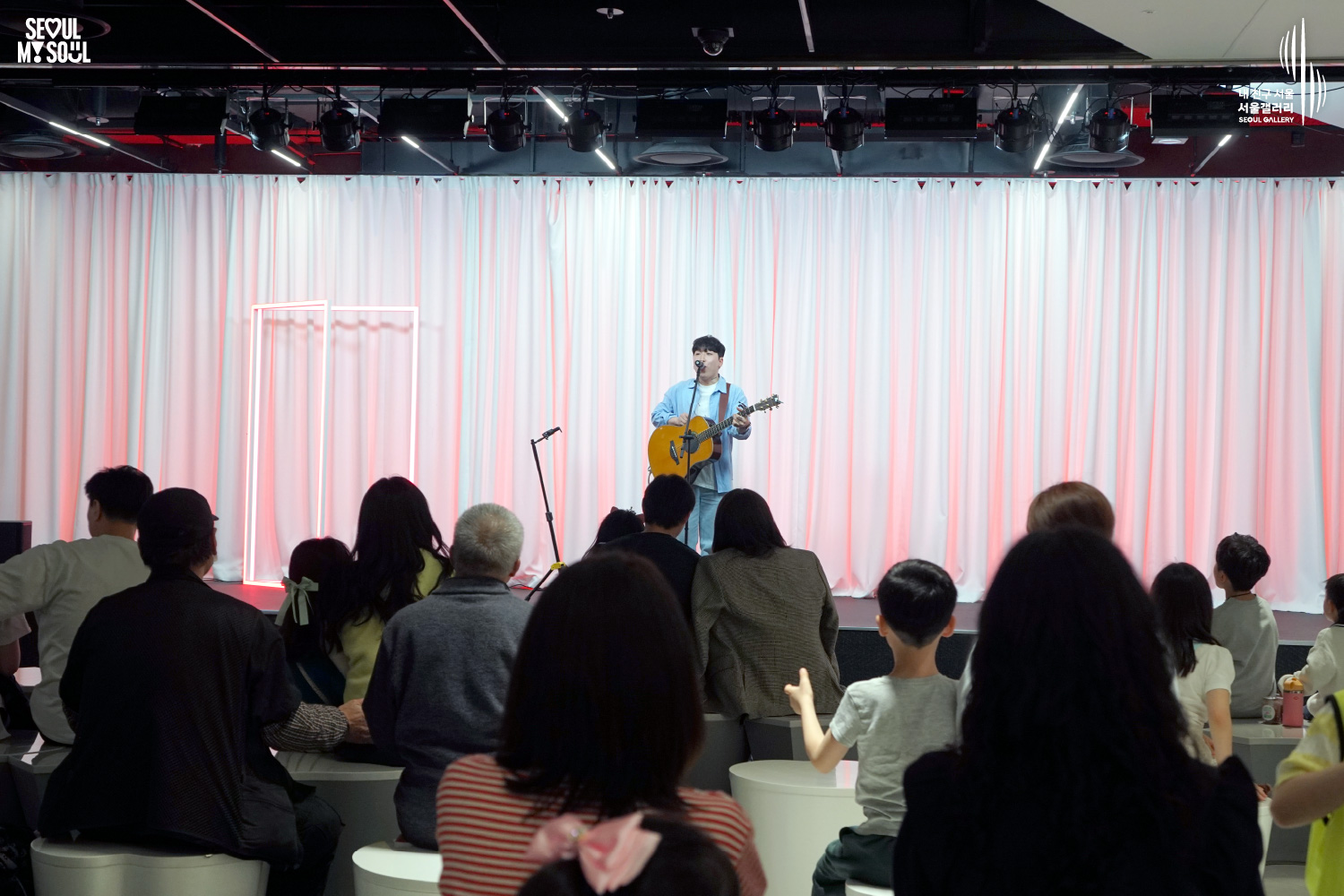 A solo acoustic guitarist performs on a pink-lit stage with white curtains at Seoul Gallery before a seated audience of all ages, with "SEOUL MY SOUL" and "Seoul Gallery" logos visible.