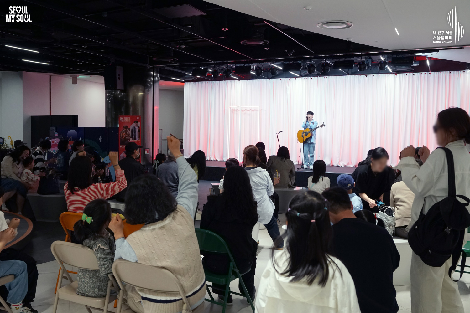 Singer-songwriter HEDYY performs with an acoustic guitar at Seoul Gallery's Lunch Stage event before a large audience of families and children, with "SEOUL MY SOUL" and "Seoul Gallery" logos visible.