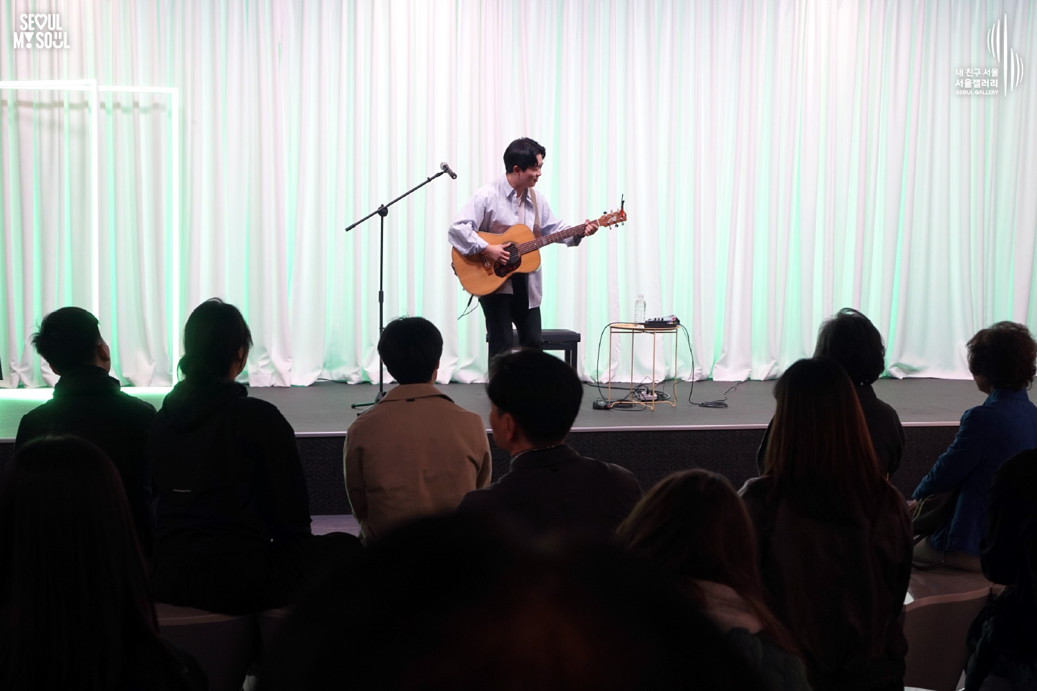 A solo acoustic guitarist performs on a green-lit stage with white curtains at Seoul Gallery before a seated audience, with "SEOUL MY SOUL" and "Seoul Gallery" logos visible.