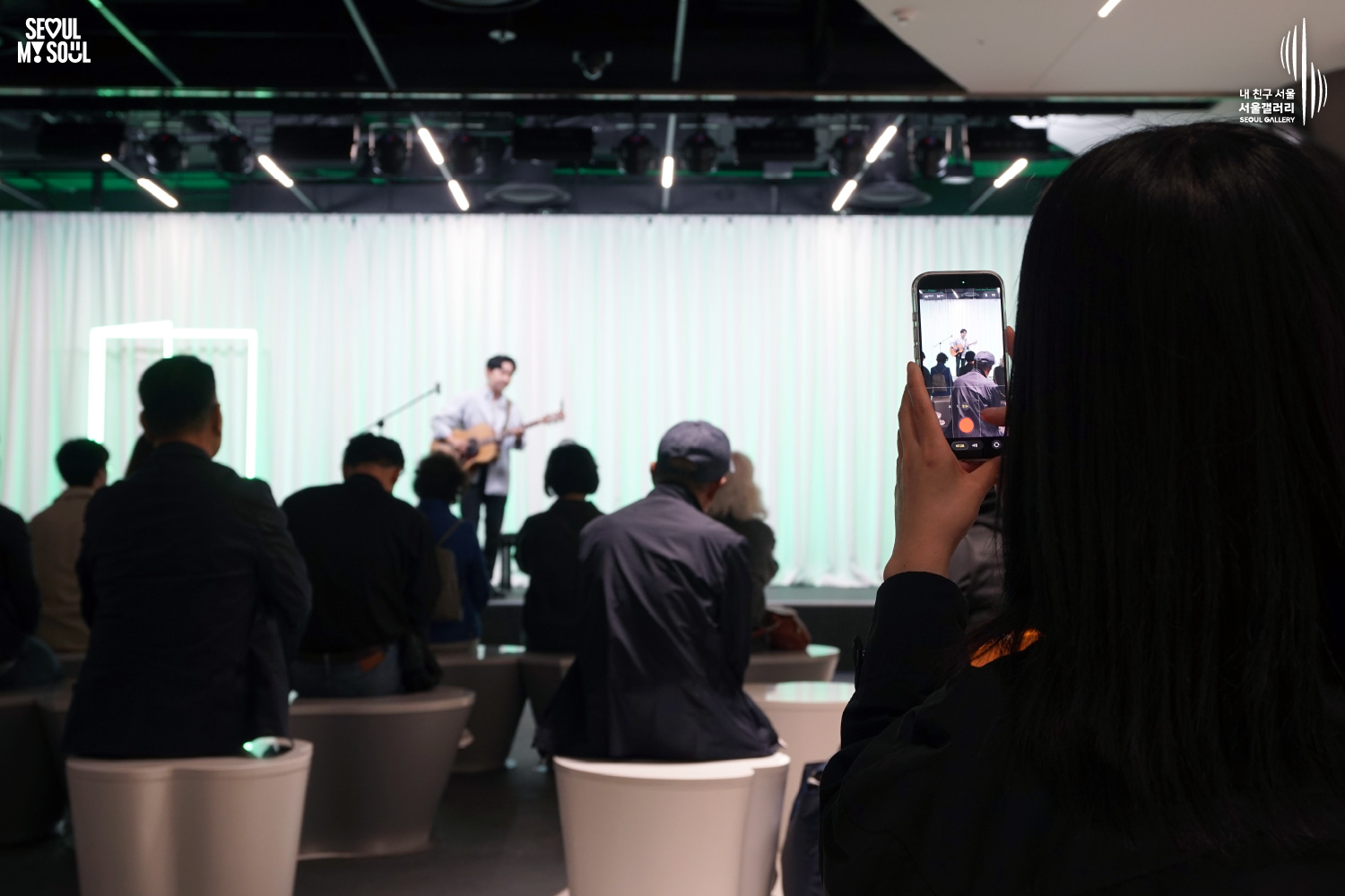An audience member films a solo acoustic guitarist's performance on a green-lit stage at Seoul Gallery, with "SEOUL MY SOUL" and "Seoul Gallery" logos visible.