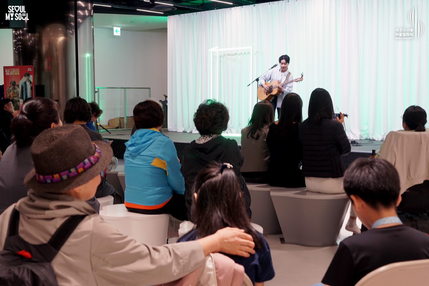 A solo acoustic guitarist performs at Seoul Gallery's Lunch Stage April event before a seated audience, with "SEOUL MY SOUL" and "Seoul Gallery" logos visible.