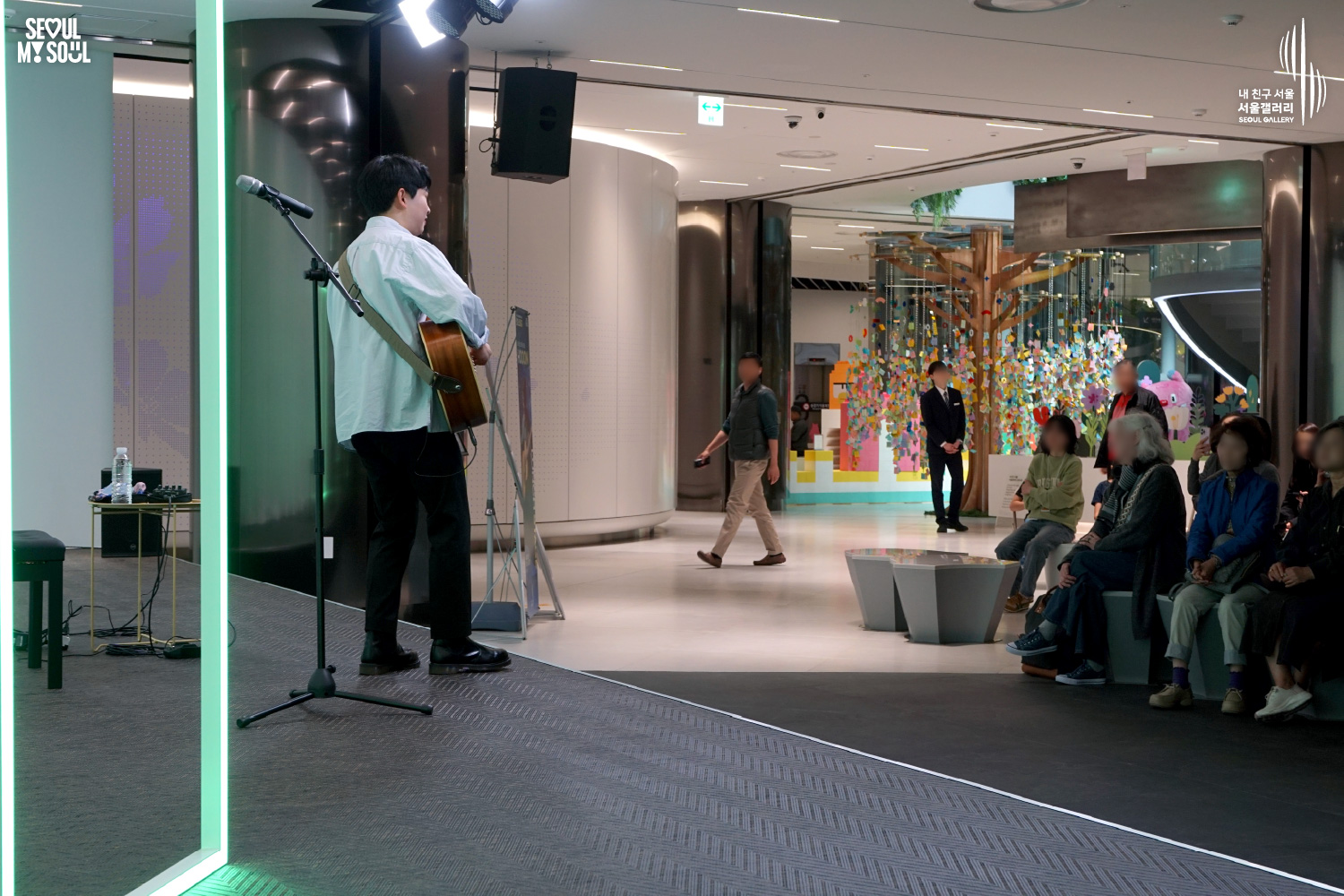 A solo acoustic guitarist performs at Seoul Gallery with a colorful tree art installation in the background and an audience watching, with "SEOUL MY SOUL" and "Seoul Gallery" logos visible.