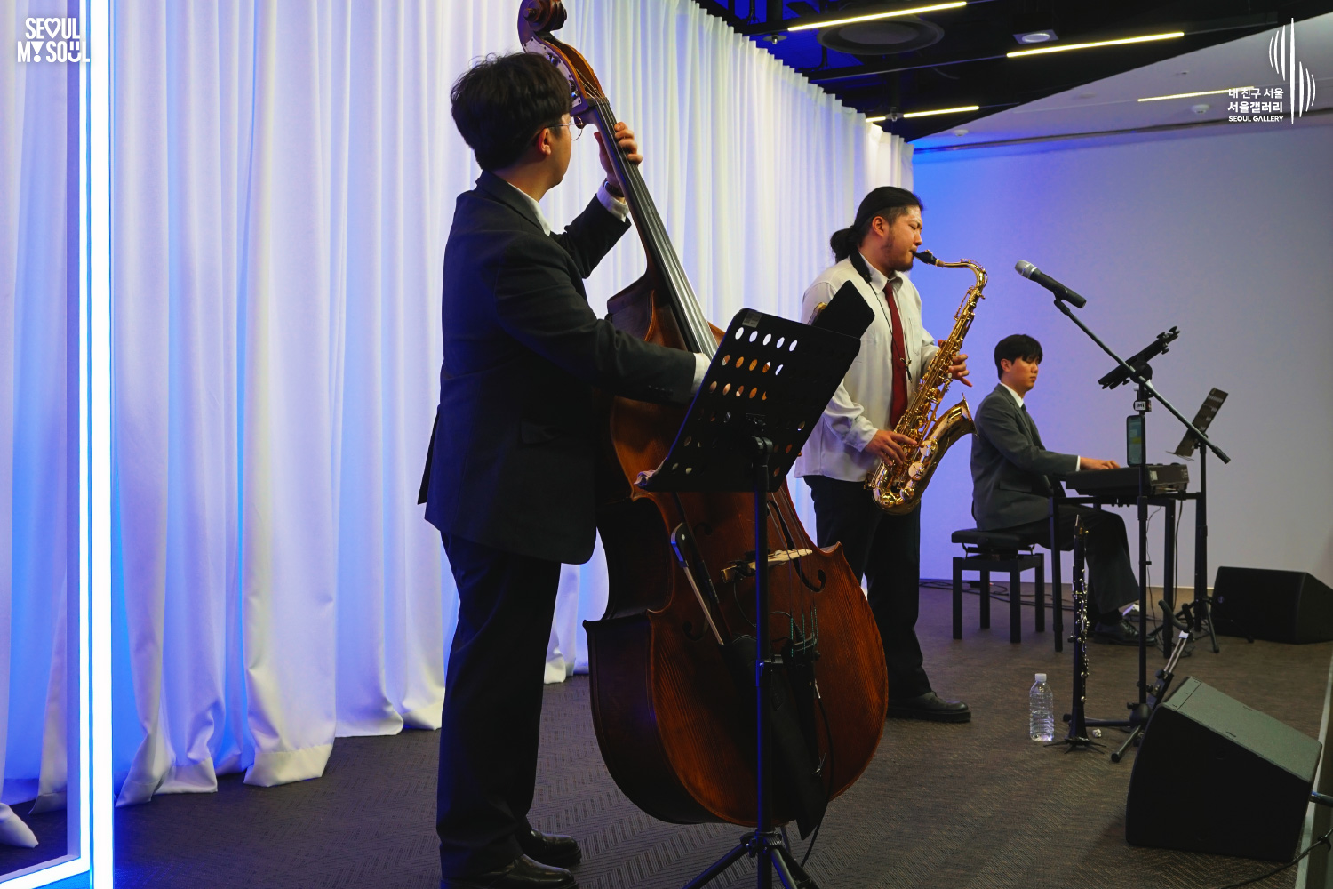 A jazz trio of double bass, saxophone, and keyboard performs on a blue-lit stage with white curtains at Seoul Gallery, with "SEOUL MY SOUL" and "Seoul Gallery" logos visible.