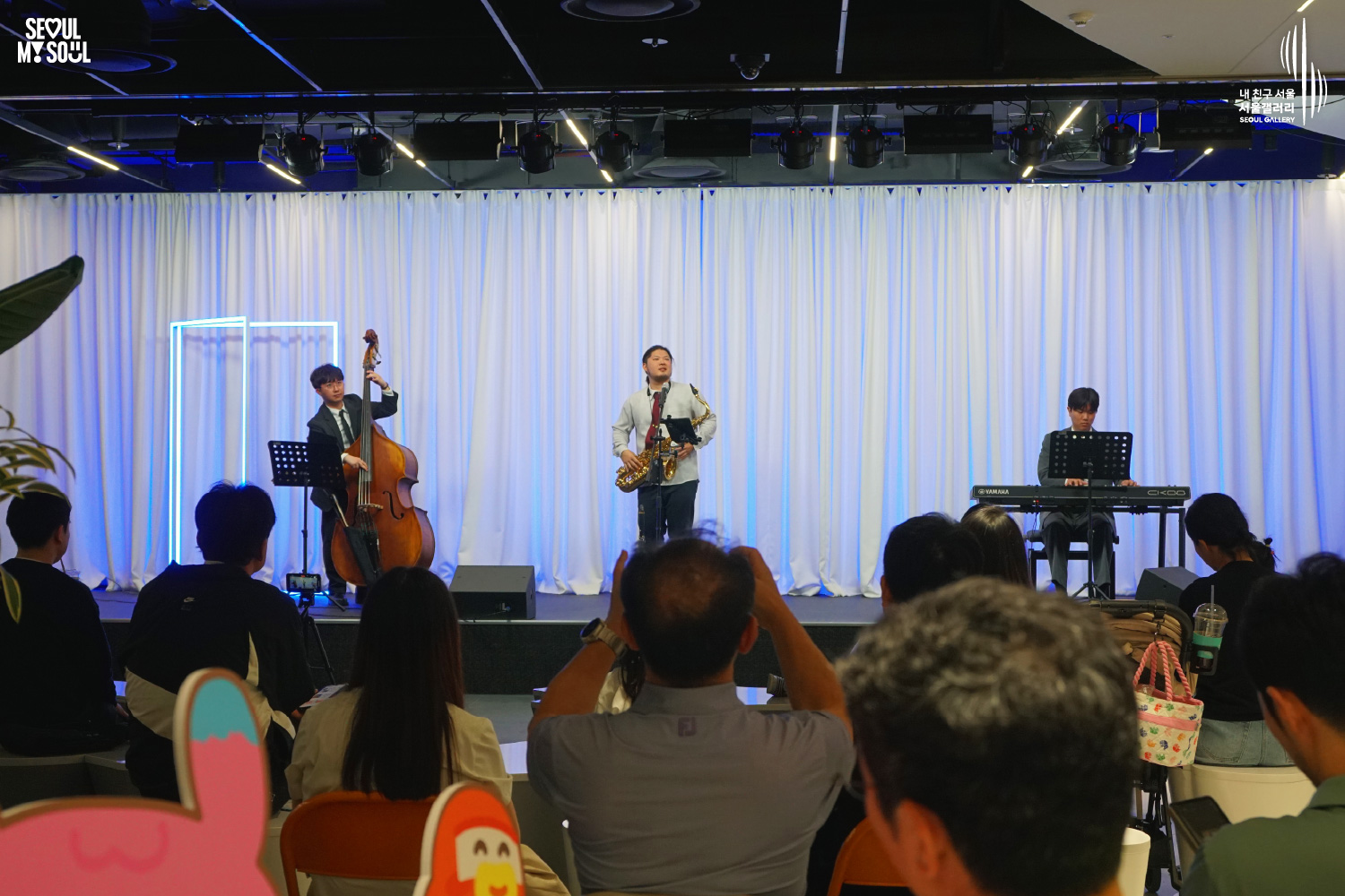 A jazz trio of double bass, saxophone, and Yamaha keyboard performs at Seoul Gallery before a seated audience, with "SEOUL MY SOUL" and "Seoul Gallery" logos visible.
