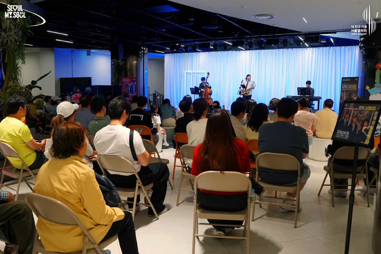 The Kim Luka Jazz Band (double bass, saxophone, keyboard) performs at Seoul Gallery's Lunch Stage event on April 25, 2026, before a seated audience, with "SEOUL MY SOUL" and "Seoul Gallery" logos visible.