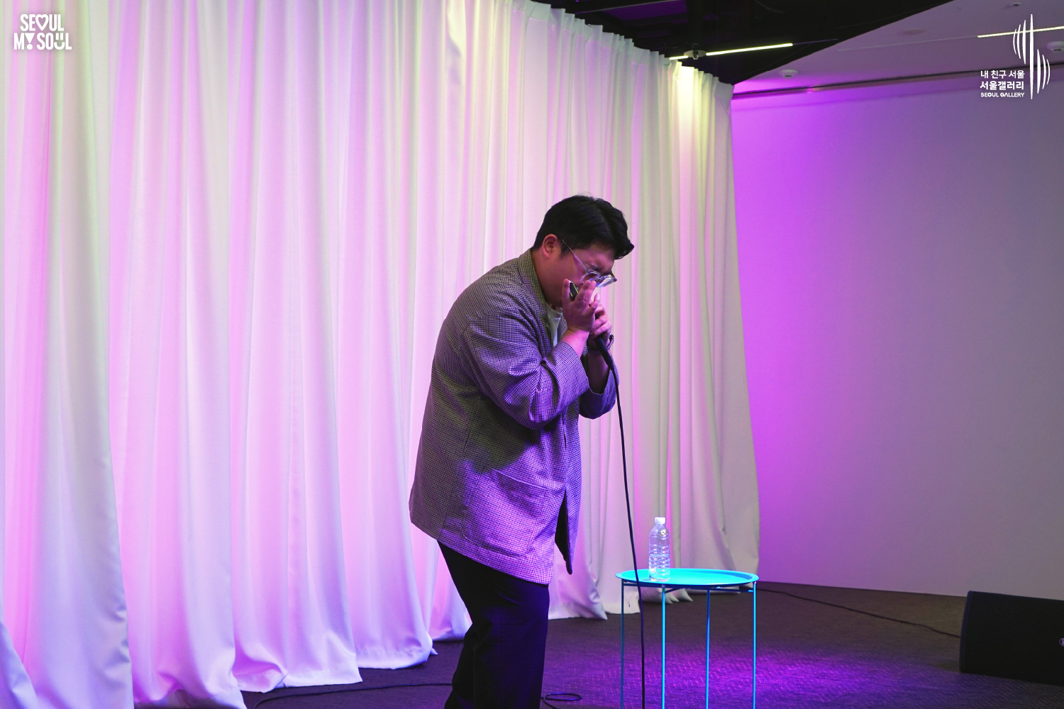 A performer in a checkered jacket speaks into a microphone on a purple-lit stage with white curtains at Seoul Gallery, with "SEOUL MY SOUL" and "Seoul Gallery" logos visible.