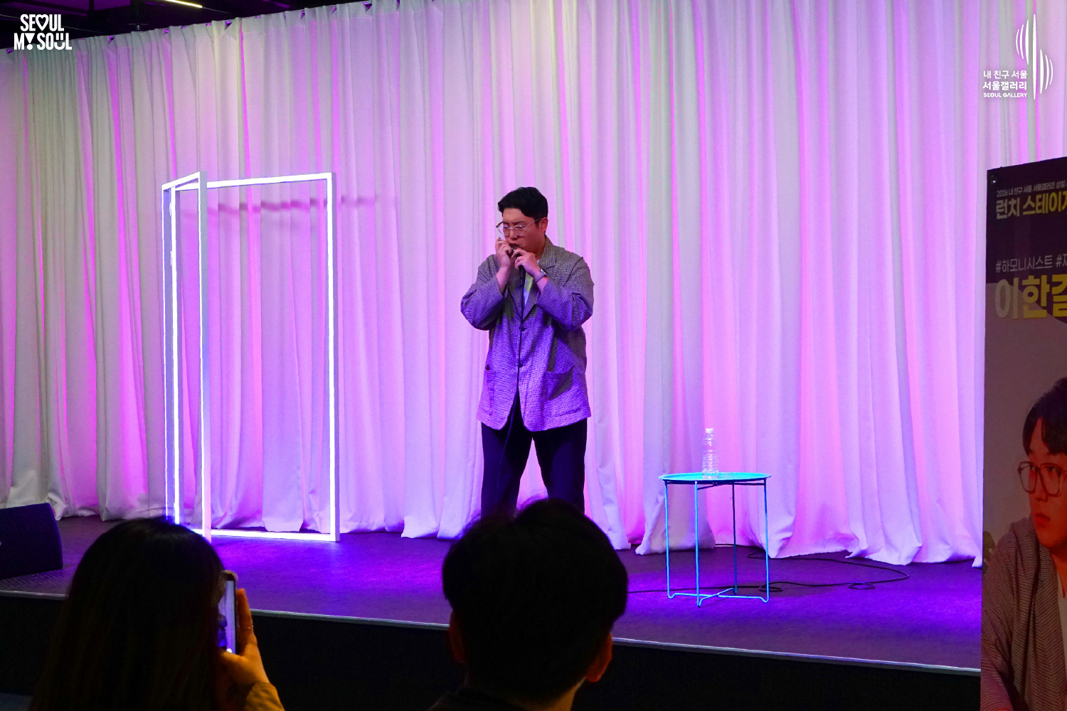 Harmonicist Lee Hangyeol plays the harmonica on a purple-lit stage during Seoul Gallery's Lunch Stage event, with audience members watching and "SEOUL MY SOUL" and "Seoul Gallery" logos visible.