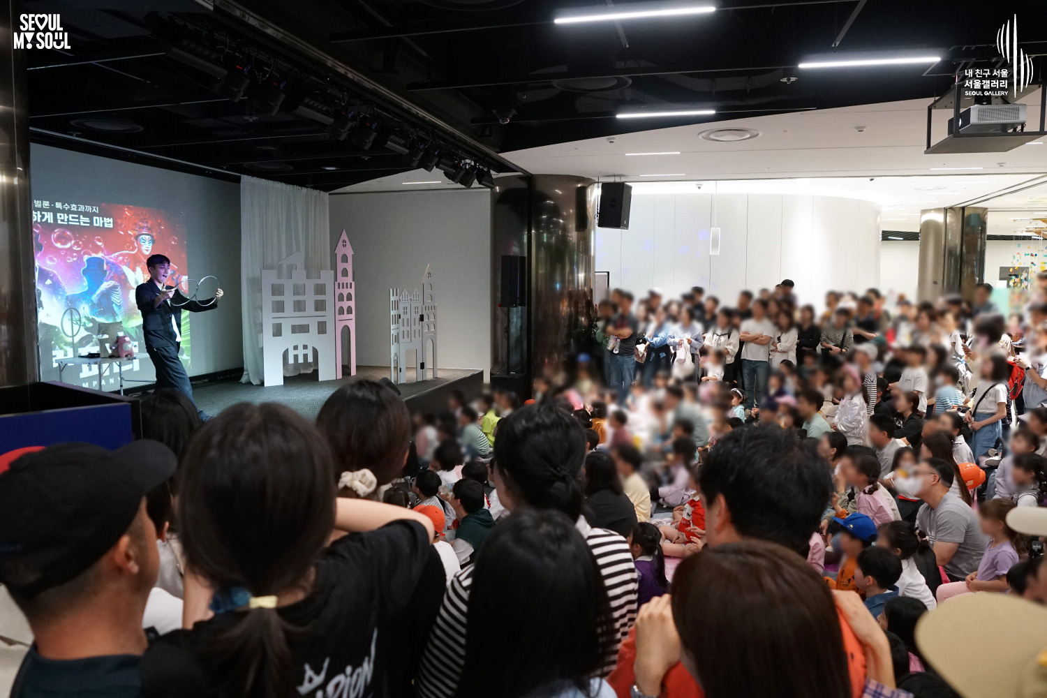 A magician performs a bubble and balloon show on the Seoul Gallery stage with decorative castle props, while a large crowd of children and families fills the venue to watch.