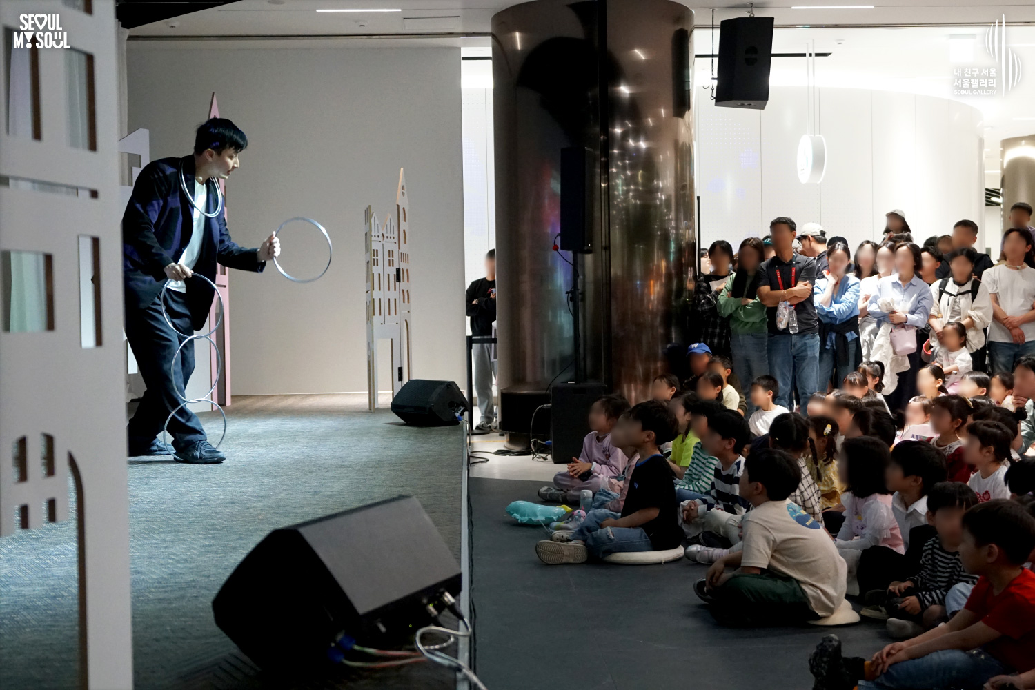 A magician performs a linking rings trick on the Seoul Gallery stage as children seated on the floor and adults standing behind watch the show.