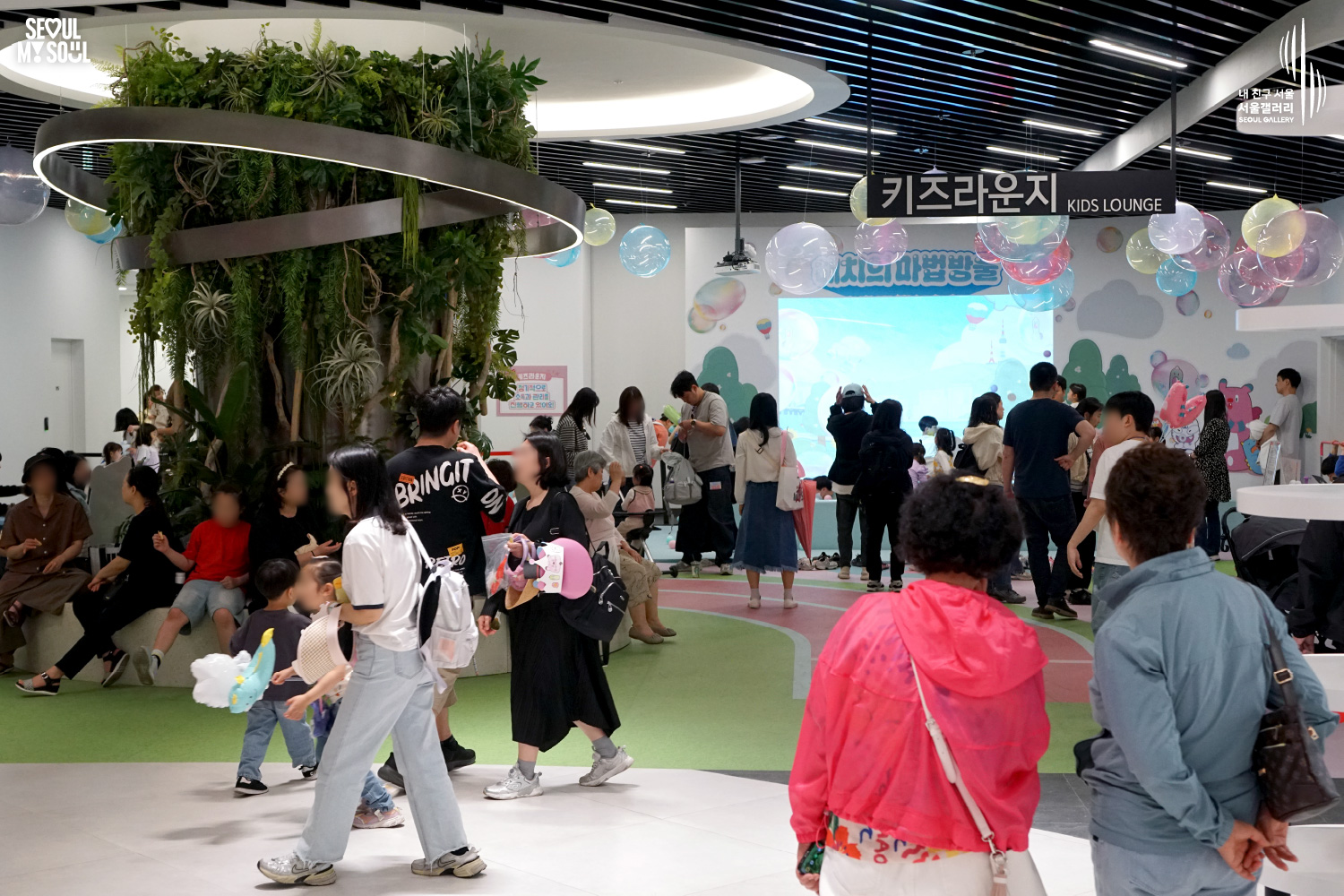 Families and children fill the Seoul Gallery Kids Lounge, exploring the colorful play area with hanging bubble decorations, plants, and a screen displaying the Hechi-themed bubble program.