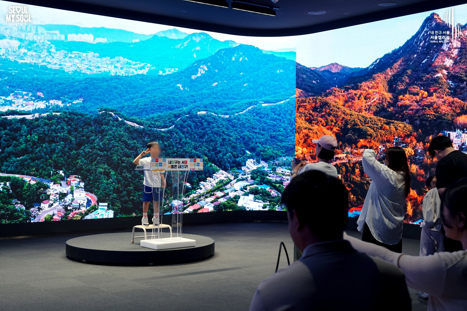 A child stands at a podium in the Seoul Gallery photo zone making a peace sign while parents take photos, with large screens displaying mountain and city landscapes of Seoul in the background.