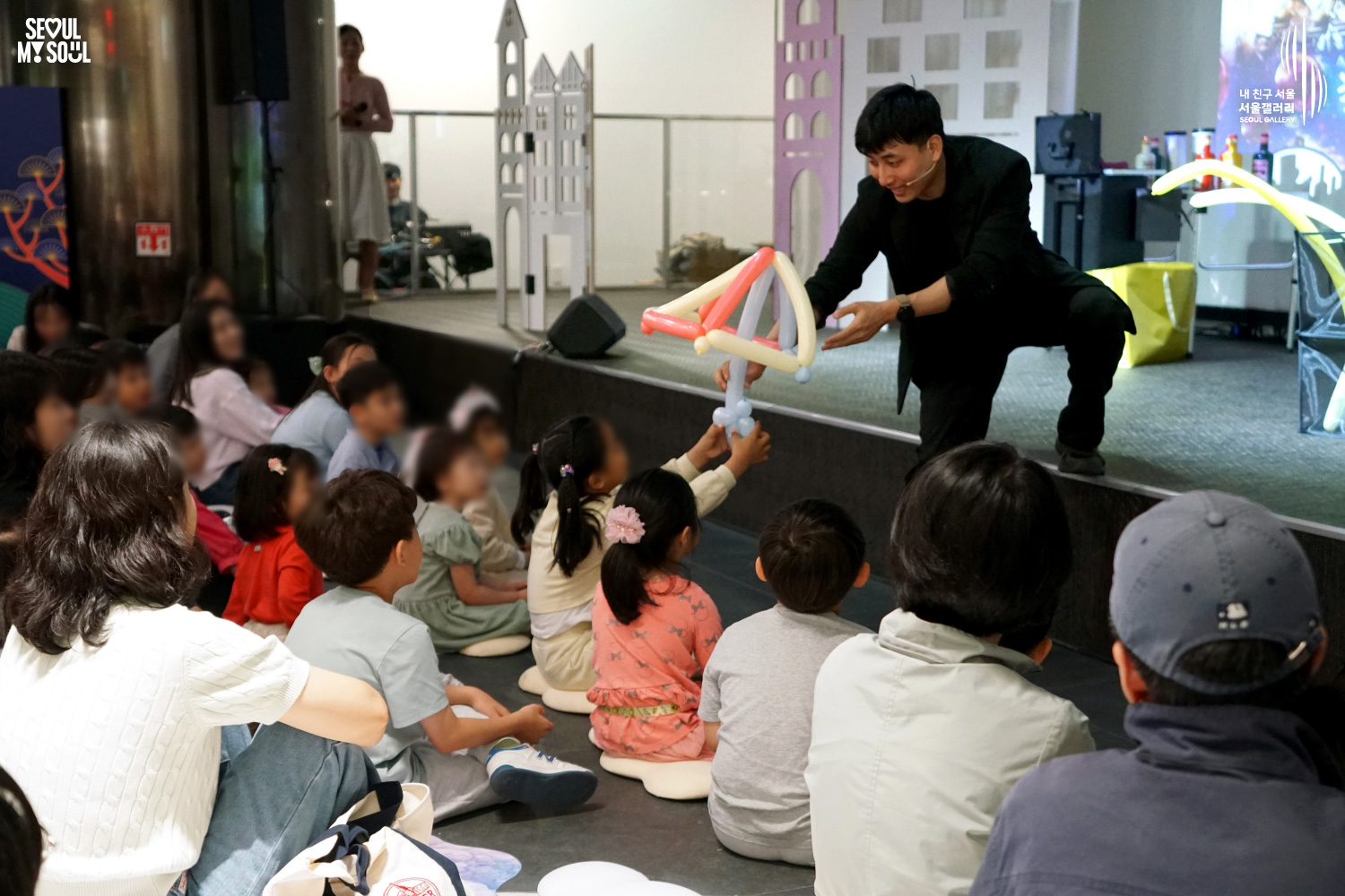 A balloon artist hands a balloon sculpture to a child during a performance on the Seoul Gallery stage, with children seated on the floor watching attentively.