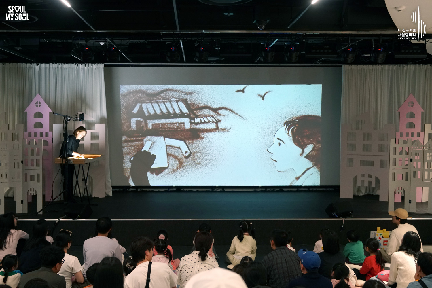 A sand artist performs on the Seoul Gallery stage with a large screen projecting her sand drawing of a traditional Korean house and a girl, as a family audience watches the show.