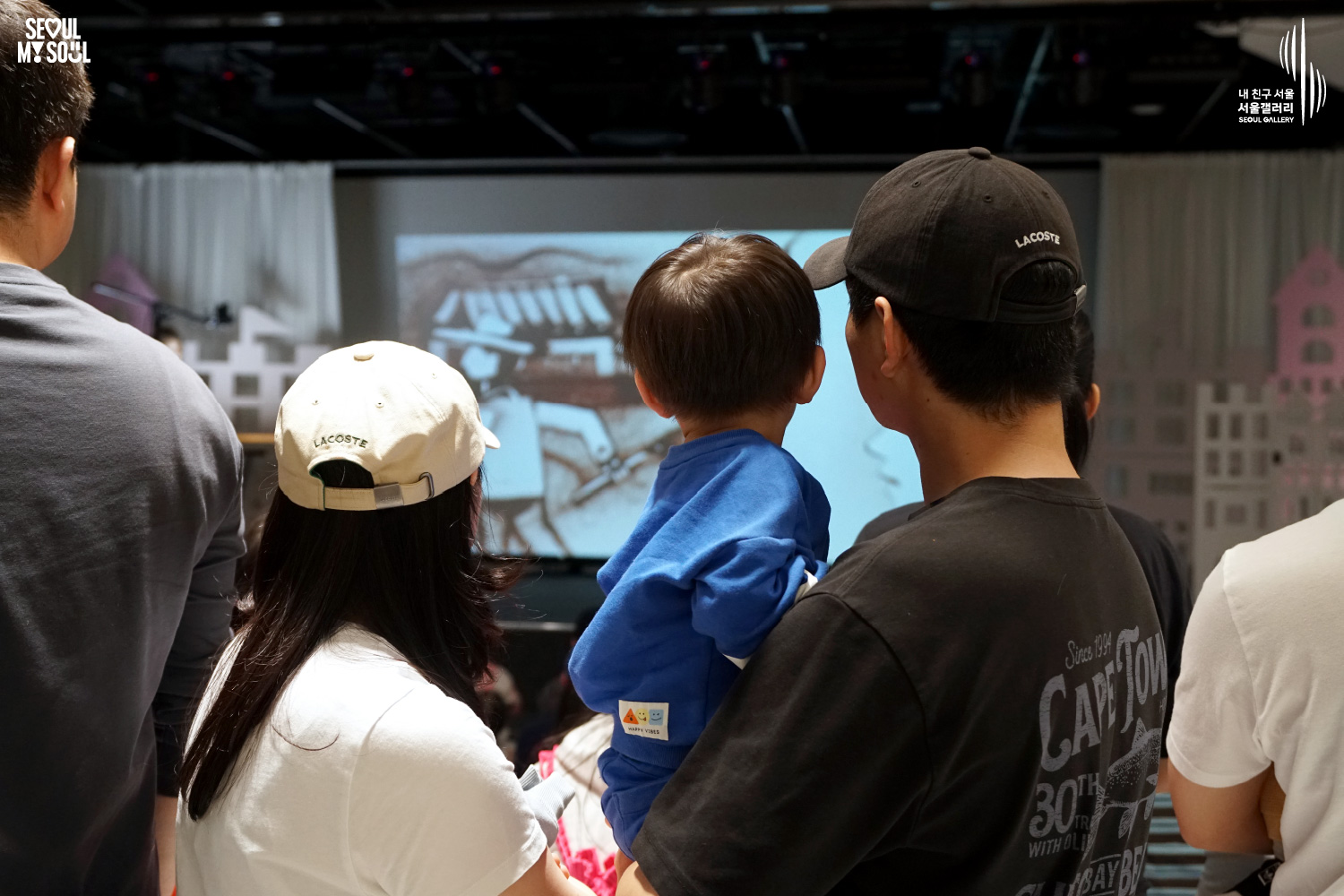A family with a young child watches a sand art performance on the Seoul Gallery stage, with the screen showing an artist's hand creating a sand drawing.