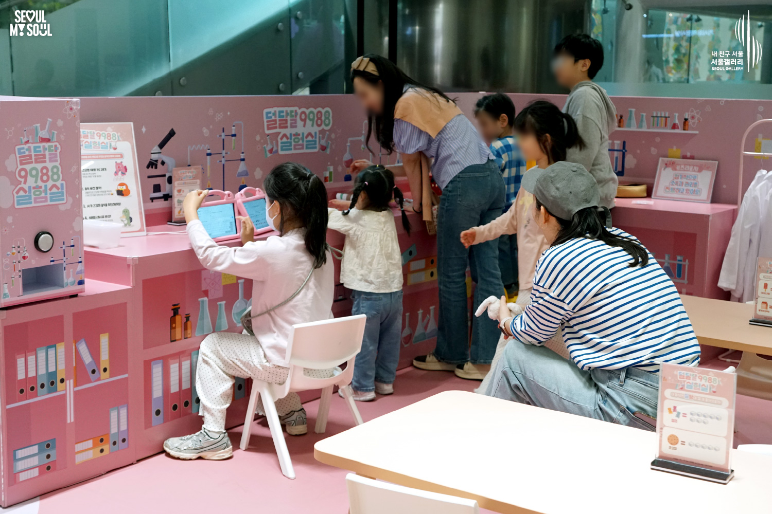 Children explore the "Deol-Dal-Dal 9988 Laboratory" experience zone at the Seoul Gallery Kids Lounge, using tablets at pink workstations while parents assist nearby in a science-themed playroom.