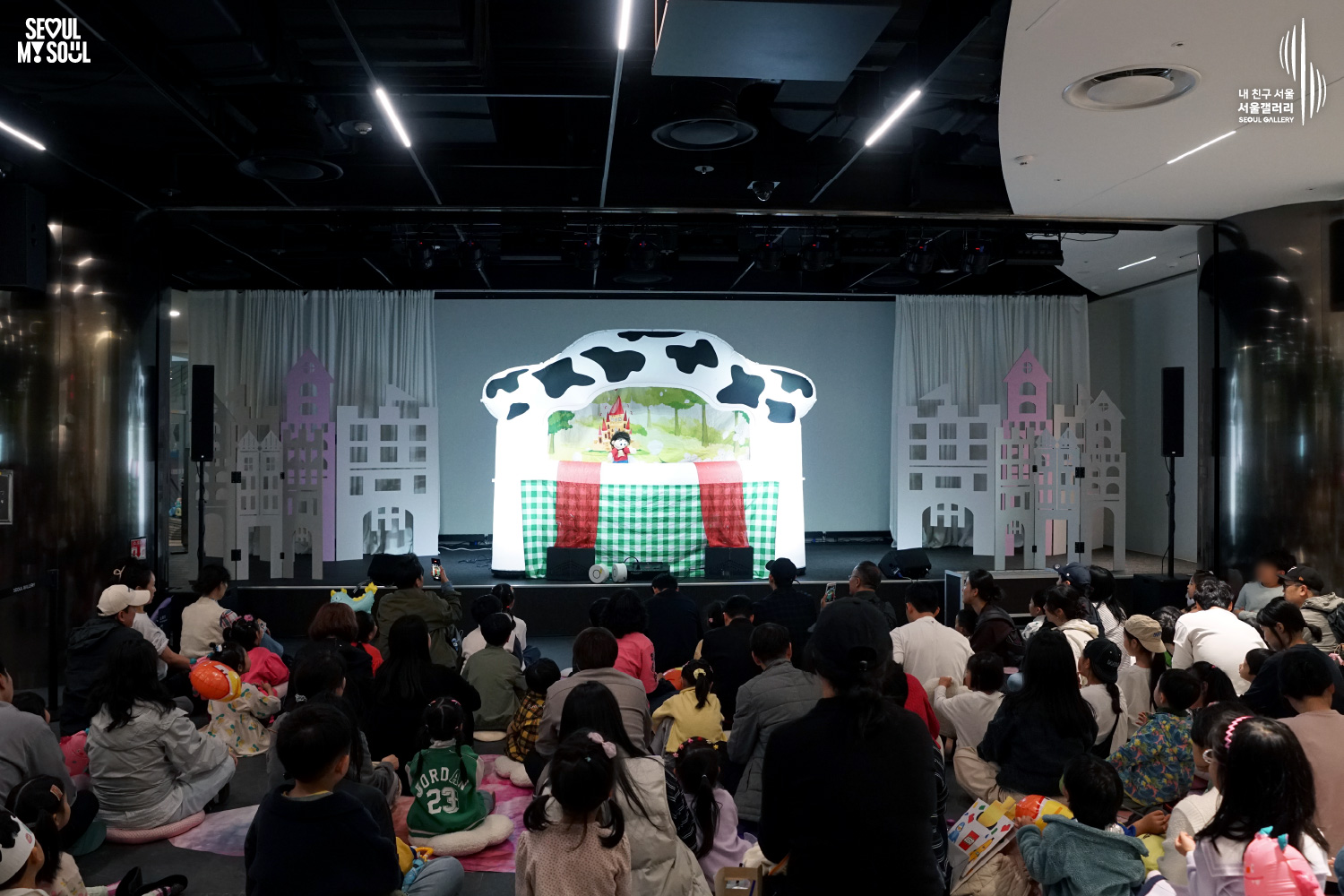 A puppet show featuring a fairytale castle scene plays on a cow-patterned puppet theater stage at Seoul Gallery, with children and families seated on the floor watching the performance.