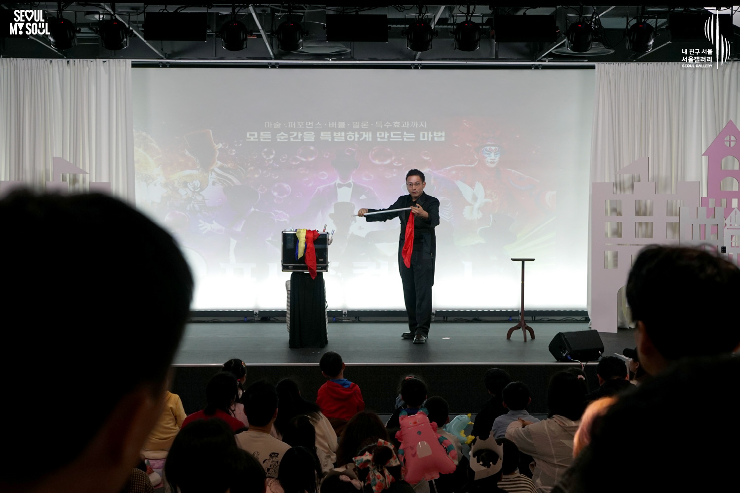 A magician performs a scarf trick on the Seoul Gallery stage with a large screen behind him advertising his magic, performance, bubble, and balloon special effects show, while an audience of children and families watches.