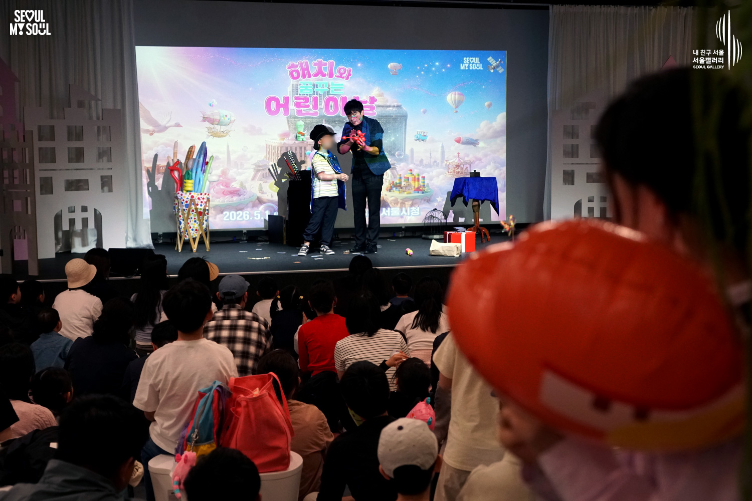 A magician performs with a young volunteer wearing a fedora on the Seoul Gallery stage at the "Children's Day with Hechi" event, with a large screen displaying the event title behind them and an audience of children watching.