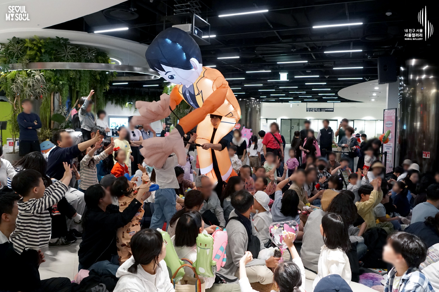 Performer in a large inflatable orange-suited character costume interacts with a seated audience of children and families at Seoul Gallery, with "SEOUL MY SOUL" and "Seoul Gallery" logos visible.