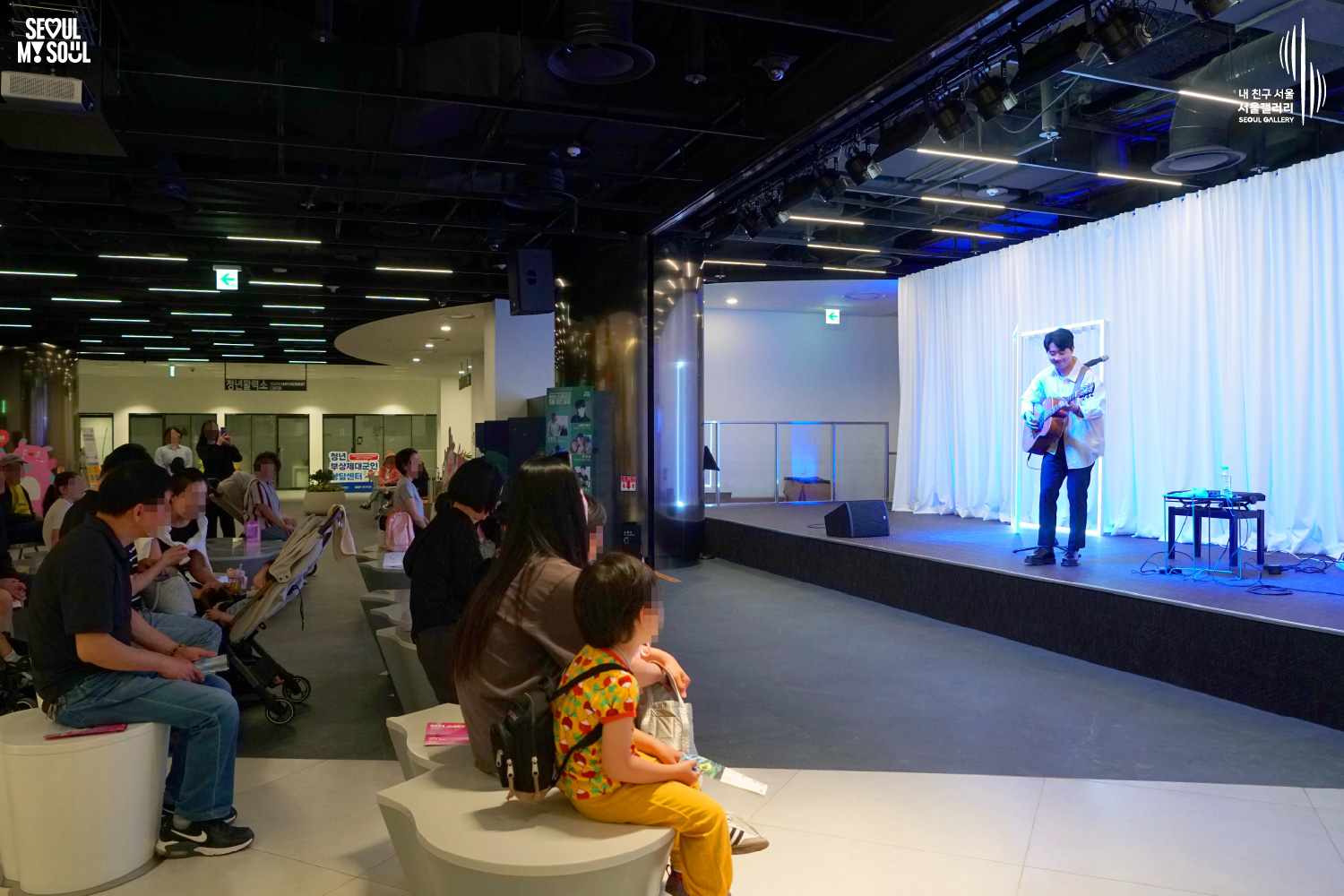 An image featuring the SEOUL MY SOUL logo and the My Friend Seoul - SEOUL GALLERY logo, capturing a live performance scene at the B1 concert hall of Seoul Gallery. On a stage with blue and square neon lighting against a white curtain background, a male singer-songwriter in a white shirt performs with an acoustic guitar. Speakers and a keyboard are arranged on the stage. In the audience, families with strollers, guardians with young children on their laps, and spectators of various ages are seated on white chairs watching the performance, while signs for the Youth Vitality Center and the Counseling Center for Discharged Wounded Soldiers are visible in the background.