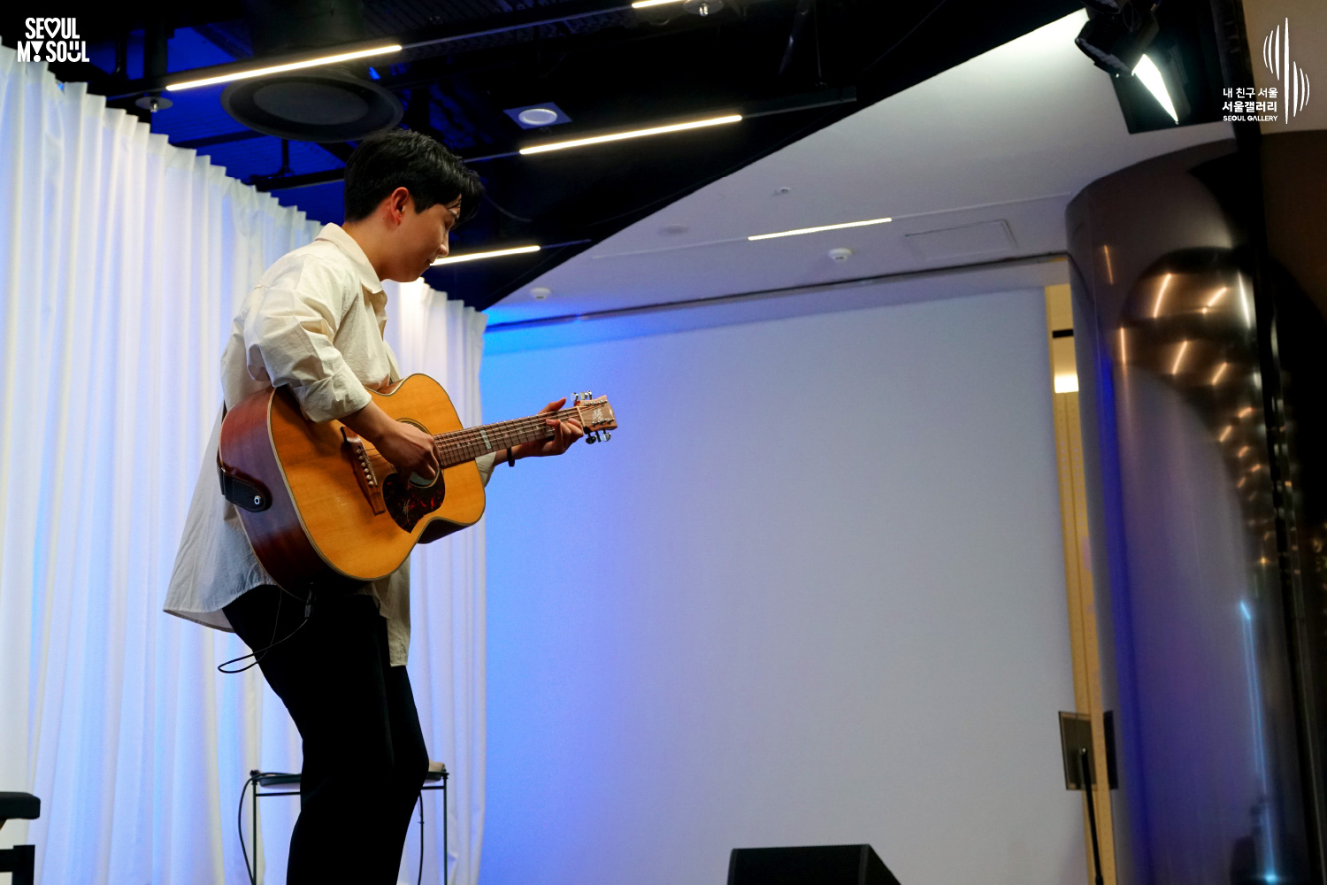 An image featuring the SEOUL MY SOUL logo and the My Friend Seoul - SEOUL GALLERY logo, capturing a stage photo at the Seoul Gallery venue. On a stage with blue lighting and a white curtain background, a profile view shows a male singer-songwriter in a white shirt and black pants, deeply immersed in playing his acoustic guitar with his head bowed and one foot resting on a chair.