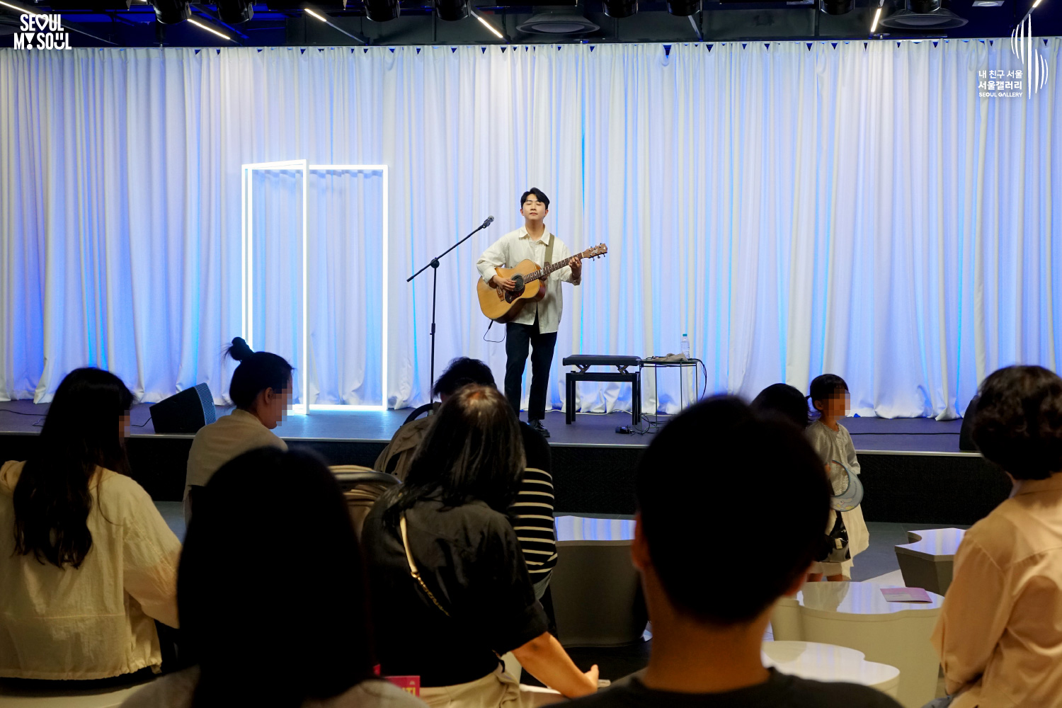 An image featuring the SEOUL MY SOUL logo and the My Friend Seoul - SEOUL GALLERY logo, capturing a live performance scene at the Seoul Gallery venue. On a stage set against a white curtain background illuminated by blue and square neon lights, a male singer-songwriter wearing a white shirt and dark blue pants sings in front of a microphone with an acoustic guitar. A chair and pedal effectors are placed on the stage, while an audience of various ages sits watching the show, and a young child stands next to the stage gazing at the performance.
