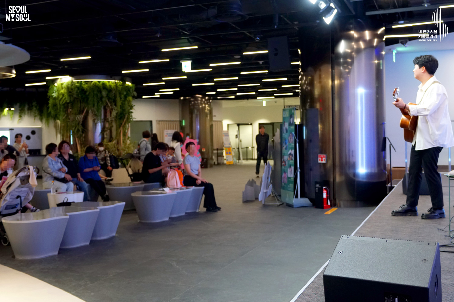 An image featuring the SEOUL MY SOUL logo and the My Friend Seoul - SEOUL GALLERY logo, capturing a live performance scene at the B1 concert hall of Seoul Gallery. On a stage within an indoor space adorned with lush green plants on one wall, a male singer-songwriter dressed in a white shirt and black pants performs with an acoustic guitar. Speakers are placed below the stage, and the audience, including families with young children and spectators of various age groups, are seated on white chairs enjoying the performance.