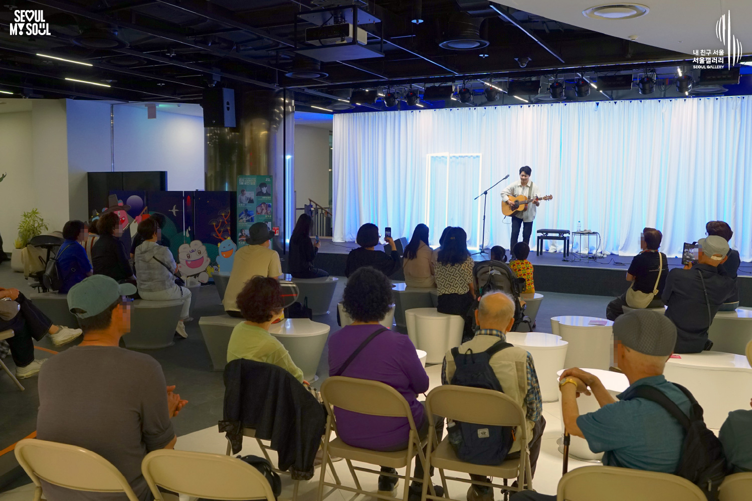 An image featuring the SEOUL MY SOUL logo and the My Friend Seoul - SEOUL GALLERY logo, capturing a live performance scene at the Seoul Gallery venue. On a stage set against a white curtain background with blended blue and square neon lighting, a male singer-songwriter in a white shirt is singing in front of a microphone with an acoustic guitar. Next to the stage, a chair, character decorations, and a performance poster are placed. In the audience, spectators of various ages are seated on white and folding chairs watching the performance, with some capturing the stage on their mobile phones.