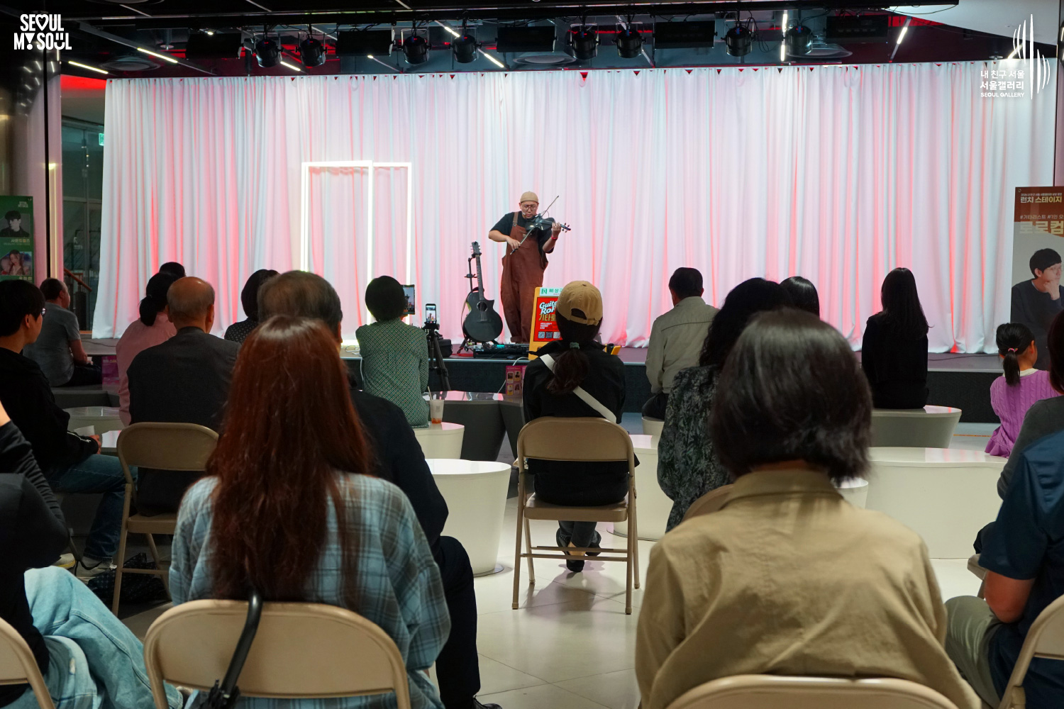 An image featuring the SEOUL MY SOUL logo and the My Friend Seoul - SEOUL GALLERY logo, capturing a live performance scene at the Seoul Gallery venue. On a stage with stage lights installed on the ceiling and pink lighting casting a glow over a white curtain background, the artist 'Guitar RoRo', wearing a beanie and bright orange overalls, plays an electric violin. Next to the stage, an acoustic guitar and a 'Guitar RoRo' sign are placed, with performance posters set up on both sides. In the audience area, spectators sit on chairs, focused intently on the stage as they enjoy the show.