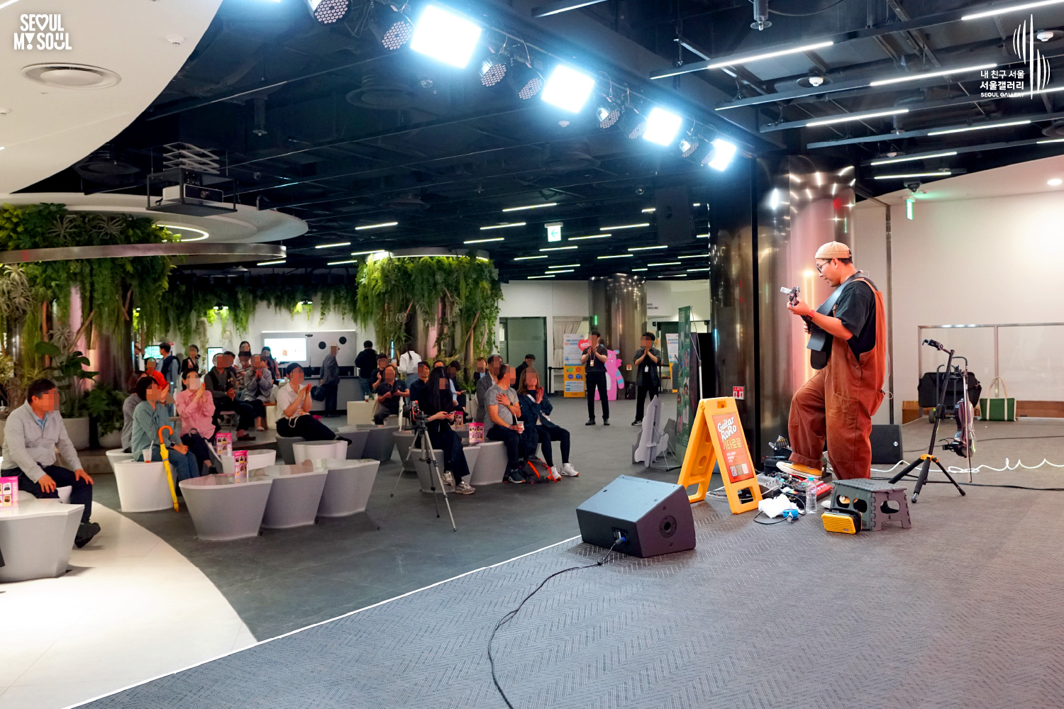 An image featuring the SEOUL MY SOUL logo and the My Friend Seoul - SEOUL GALLERY logo, capturing a live performance scene at the B1 concert hall of Seoul Gallery. In an indoor space where stage lights are installed on the ceiling and one wall is adorned with lush green plants, the artist 'Guitar RoRo', wearing a beanie and orange overalls, performs on stage with a guitar. In front of the stage, speakers, a microphone stand, and a 'Guitar RoRo' sign are placed, while the audience sits on white chairs arranged in a curved layout, clapping and enjoying the performance.