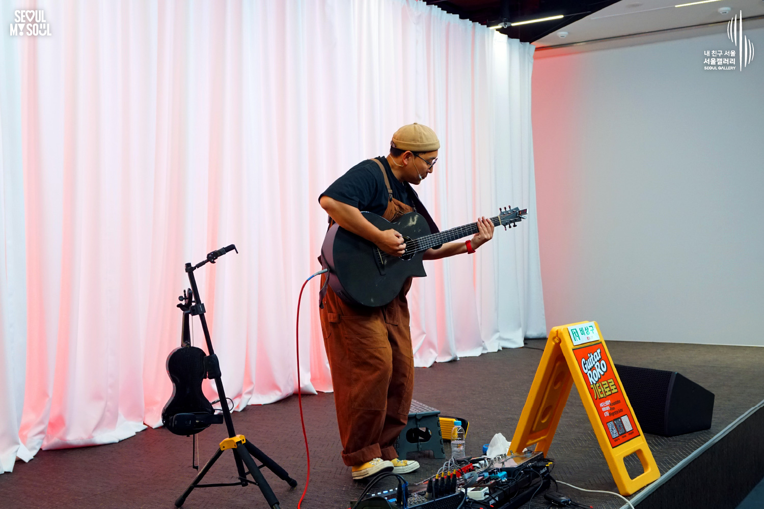 An image featuring the SEOUL MY SOUL logo and the My Friend Seoul - SEOUL GALLERY logo, capturing a stage photo at the Seoul Gallery venue. On a stage with a pink-lit white curtain background, the artist 'Guitar RoRo', wearing a beanie, glasses, a black T-shirt, and bright orange overalls, is deeply immersed in playing an acoustic guitar, holding it with both hands. Arranged on the stage are a microphone stand, an electric violin, pedal effectors, speakers, a 'Guitar RoRo' sign, and an emergency exit sign.