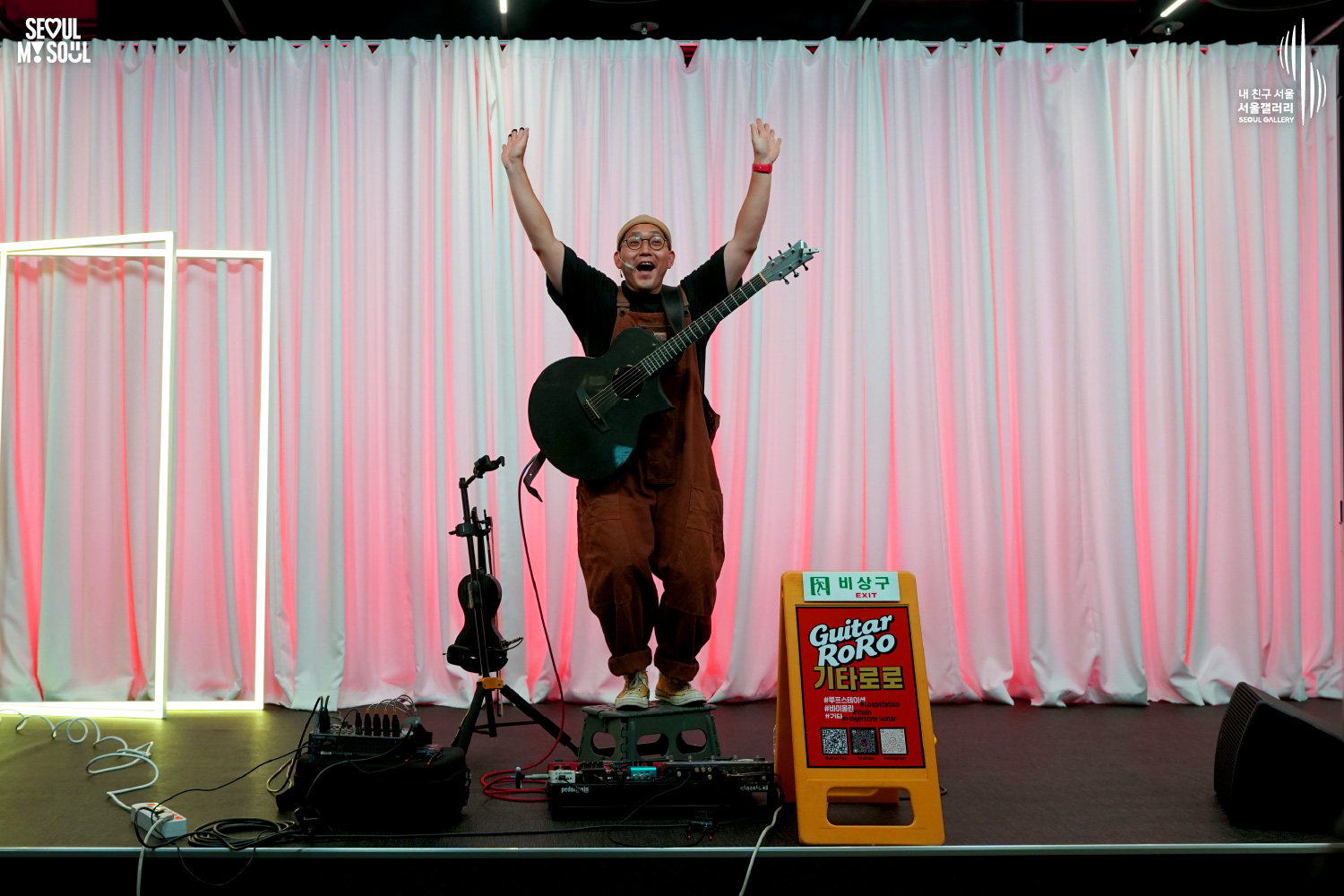 An image featuring the SEOUL MY SOUL logo and the My Friend Seoul - SEOUL GALLERY logo, capturing a stage photo at the Seoul Gallery venue. On a stage set against a white curtain background illuminated by pink lighting, the artist 'Guitar RoRo', wearing a beanie, glasses, a black T-shirt, and bright orange overalls, stands on a small footstool with both arms wide open, greeting the audience with a bright expression while carrying an acoustic guitar. The stage also features a microphone stand, pedal effectors, speakers, a 'Guitar RoRo' information sign, and an emergency exit sign.