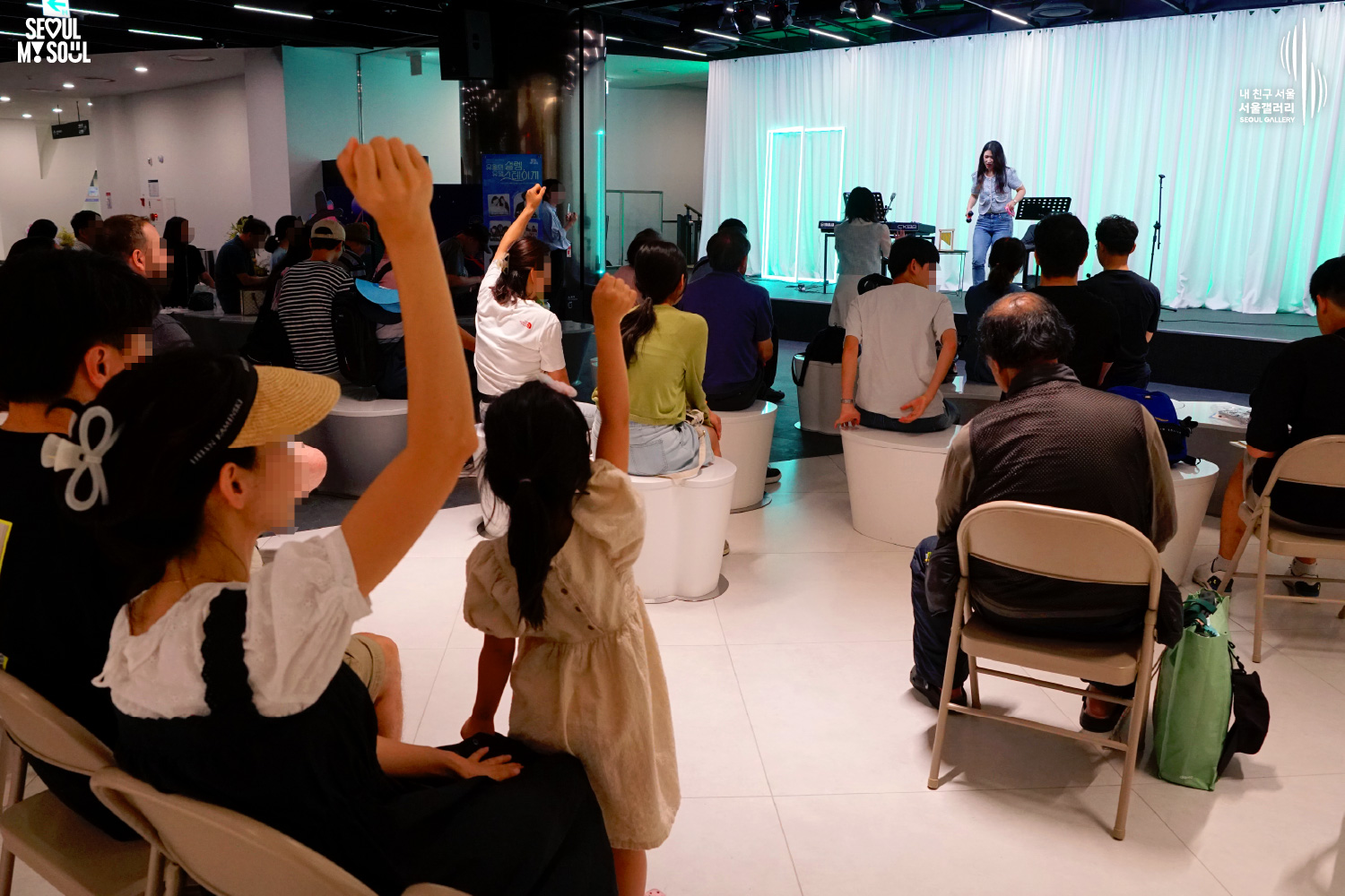SEOUL MY SOUL 로고와 내 친구 서울 서울갤러리 SEOUL GALLERY 로고가 표기된 서울갤러리 공연장 사진으로, 연둣빛 조명과 사각 네온 조명이 어우러진 흰색 커튼 배경의 무대 위에서 두 명의 여성 아티스트가 공연을 진행하고 있으며, 객석에는 다양한 연령대의 관람객들이 앉아 공연을 관람하고 있고 전경에는 어린아이들이 손을 들어 무대를 향해 반응하는 모습이 담긴 이미지입니다.