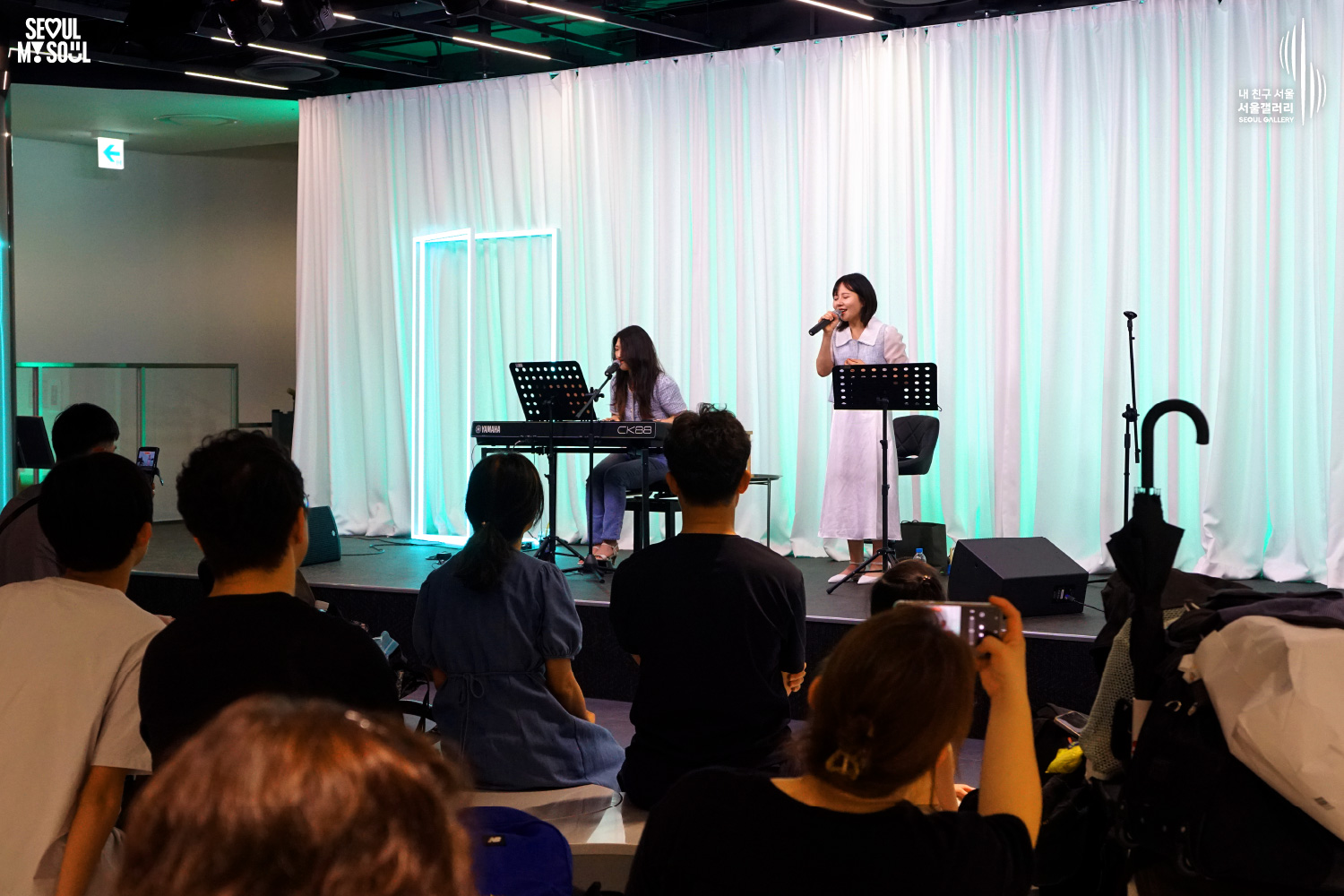 SEOUL MY SOUL 로고와 내 친구 서울 서울갤러리 SEOUL GALLERY 로고가 표기된 서울갤러리 공연장 사진으로, 연둣빛 조명과 사각 네온 조명이 어우러진 흰색 커튼 배경의 무대 위에서 여성 연주자가 키보드를 연주하고 있으며 여성 싱어송라이터가 마이크를 들고 노래를 부르고 있고, 객석에는 관람객들이 공연을 관람하며 일부는 휴대전화로 무대를 촬영하고 있는 모습이 담긴 이미지입니다.