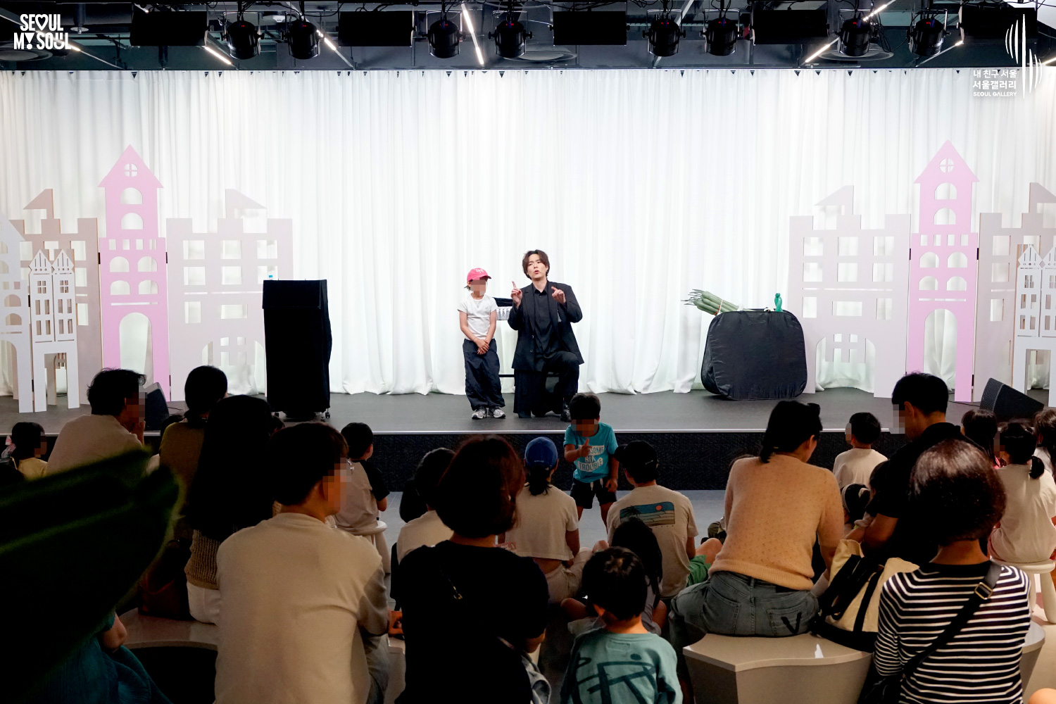 SEOUL MY SOUL 로고와 내 친구 서울 서울갤러리 SEOUL GALLERY 로고가 표기된 서울갤러리 공연장 사진으로, 무대 조명이 비추는 흰색 커튼 배경의 무대 위에서 중간 길이 머리의 재킷을 착용한 남성 아티스트가 어린이 참여자와 함께 공연을 진행하고 있으며, 무대 위에는 상자와 테이블이 놓여 있고 객석에는 어린아이를 동반한 가족과 관람객들이 무대를 바라보고 있는 모습이 담긴 이미지입니다.