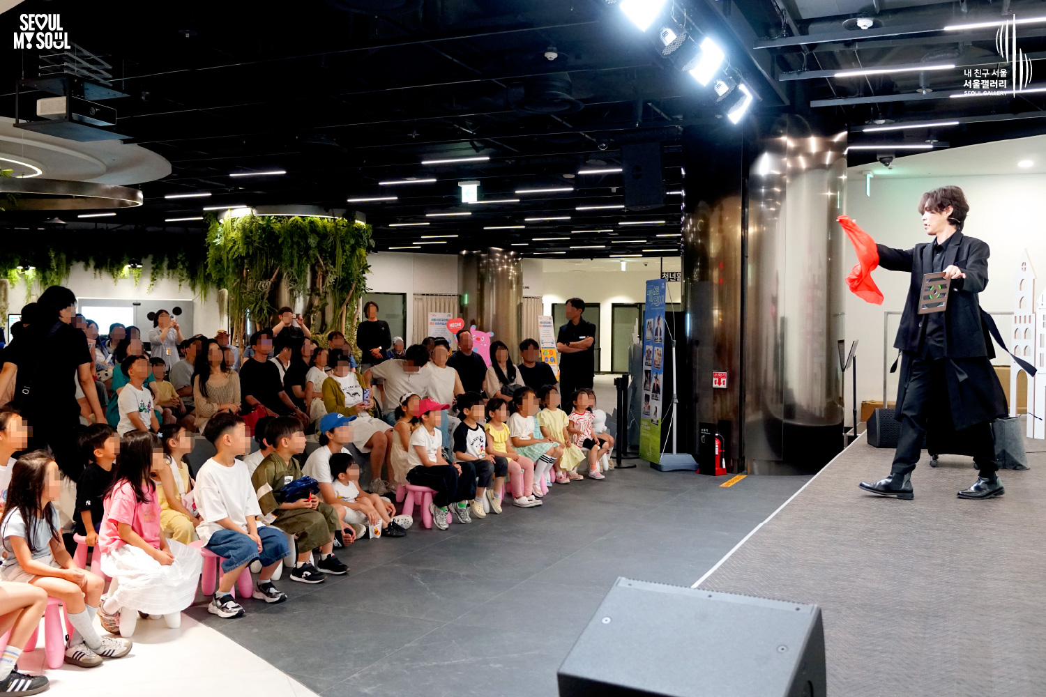 SEOUL MY SOUL 로고와 내 친구 서울 서울갤러리 SEOUL GALLERY 로고가 표기된 서울갤러리 지하 1층 공연장의 공연 현장 사진으로, 천장에 무대 조명이 설치된 실내 공간에서 중간 길이 머리의 재킷을 착용한 남성 아티스트가 천을 활용한 마술을 선보이고 있으며, 객석 앞줄에는 어린이 관람객들이 앉아 무대를 바라보고 있고 뒤편에는 가족 단위 관람객들이 공연을 관람하고 있는 모습이 담긴 이미지입니다.