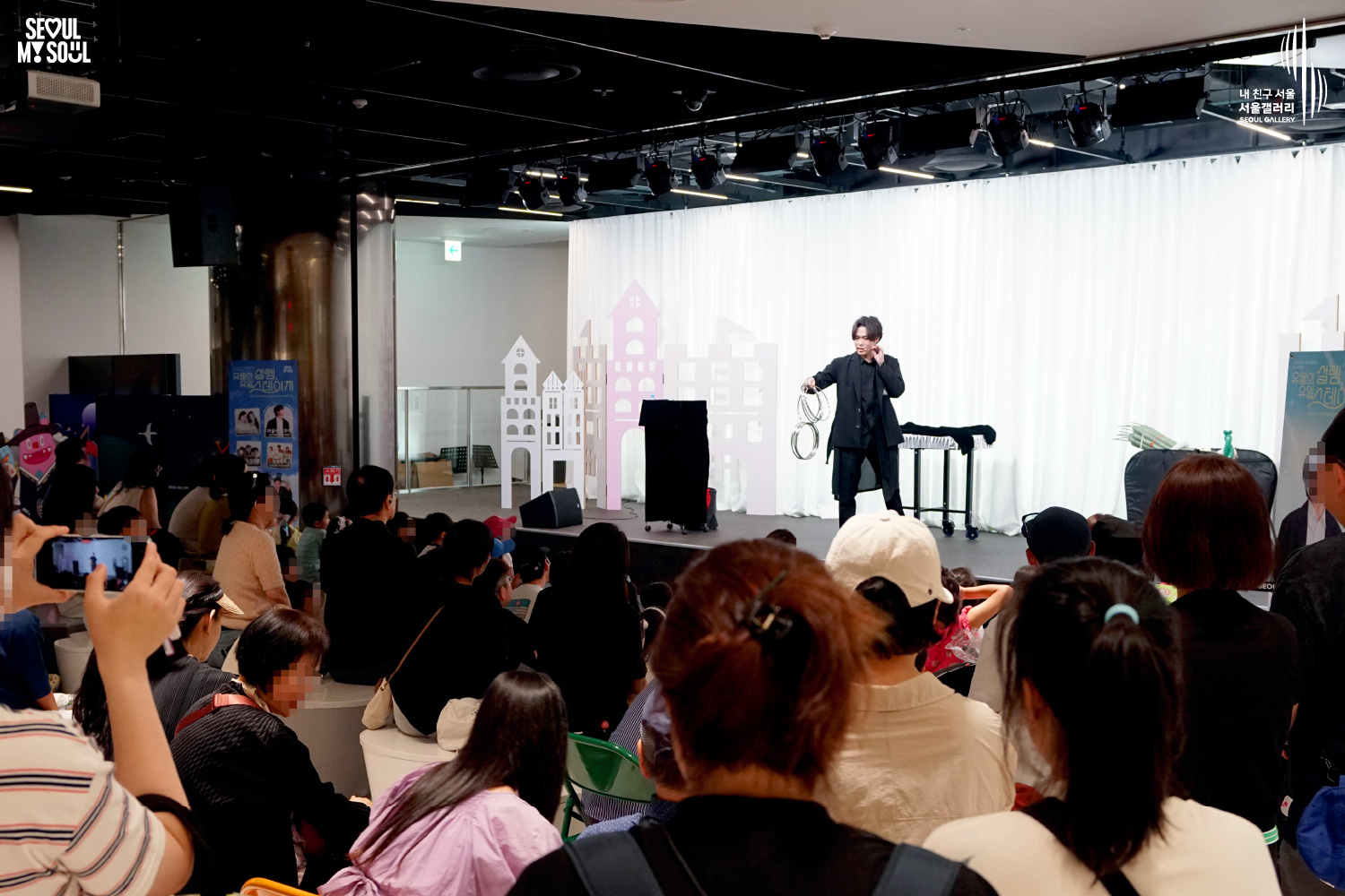 SEOUL MY SOUL 로고와 내 친구 서울 서울갤러리 SEOUL GALLERY 로고가 표기된 서울갤러리 공연장 사진으로, 무대 조명이 비추는 흰색 커튼 배경의 무대 위에서 중간 길이 머리의 재킷을 착용한 남성 아티스트가 고리 형태의 공연 소품을 들고 공연을 진행하고 있으며, 무대 위에는 테이블과 상자, 공연 소품이 놓여 있고 객석에는 관람객들이 공연을 관람하며 일부는 휴대전화로 무대를 촬영하고 있는 모습이 담긴 이미지입니다.