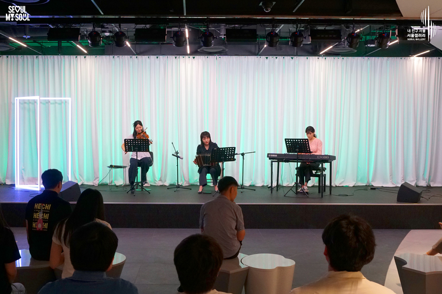 SEOUL MY SOUL 로고와 내 친구 서울 서울갤러리 SEOUL GALLERY 로고가 표기된 서울갤러리 공연장 사진으로, 푸른빛 조명과 사각 네온 조명이 어우러진 흰색 커튼 배경의 무대 위에서 여성 연주자 세 명이 바이올린과 반도네온, 키보드를 연주하고 있으며, 무대 양쪽에는 모니터 스피커가 놓여 있고 객석에는 관람객들이 앉아 공연을 관람하고 있는 모습이 담긴 이미지입니다.